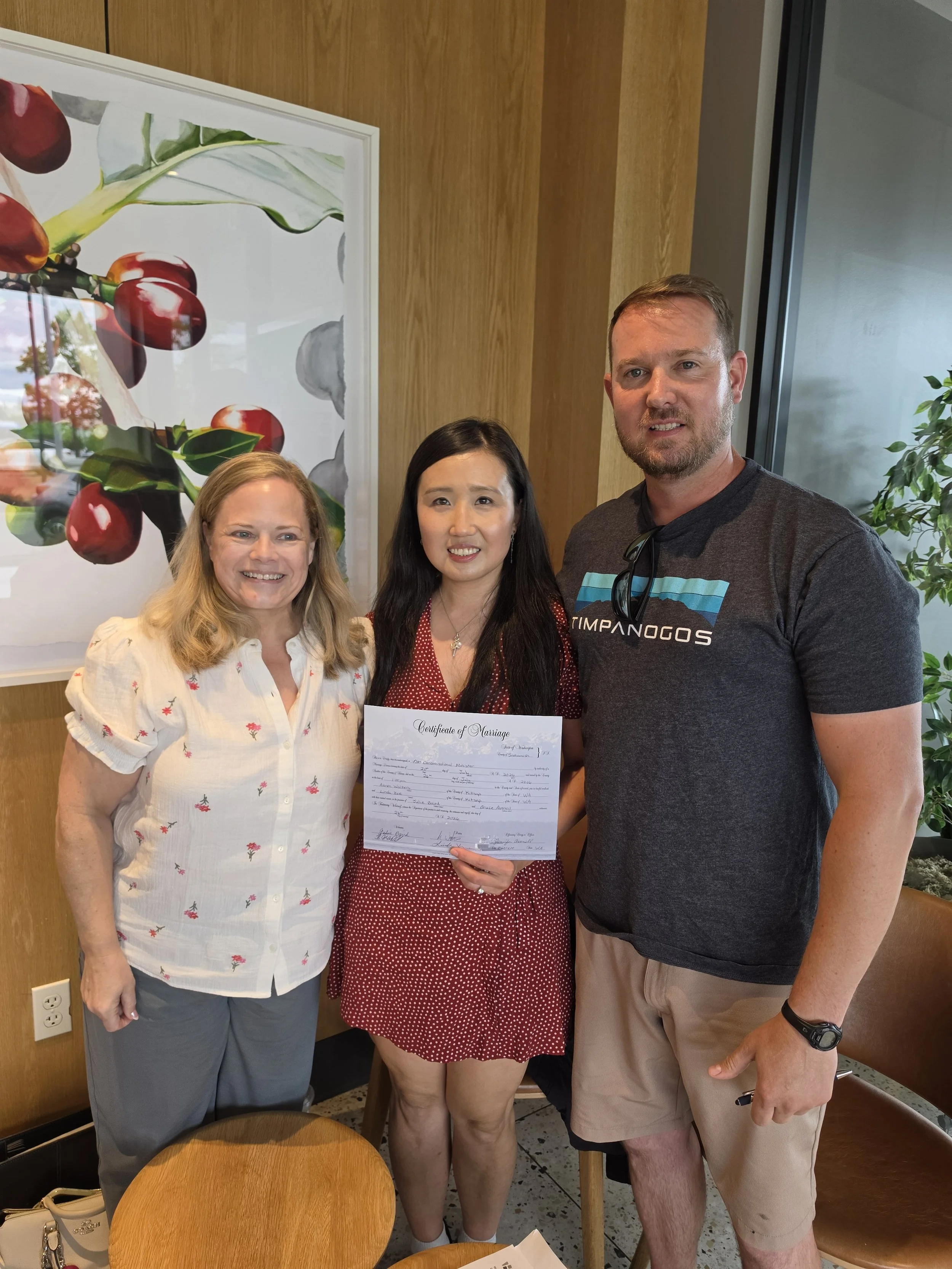 Three people standing together, two women and one man, smiling indoors, with one woman holding a marriage certificate. They are near a wooden wall with a large framed artwork of coffee cherries on the left and a window with a plant on the right.
