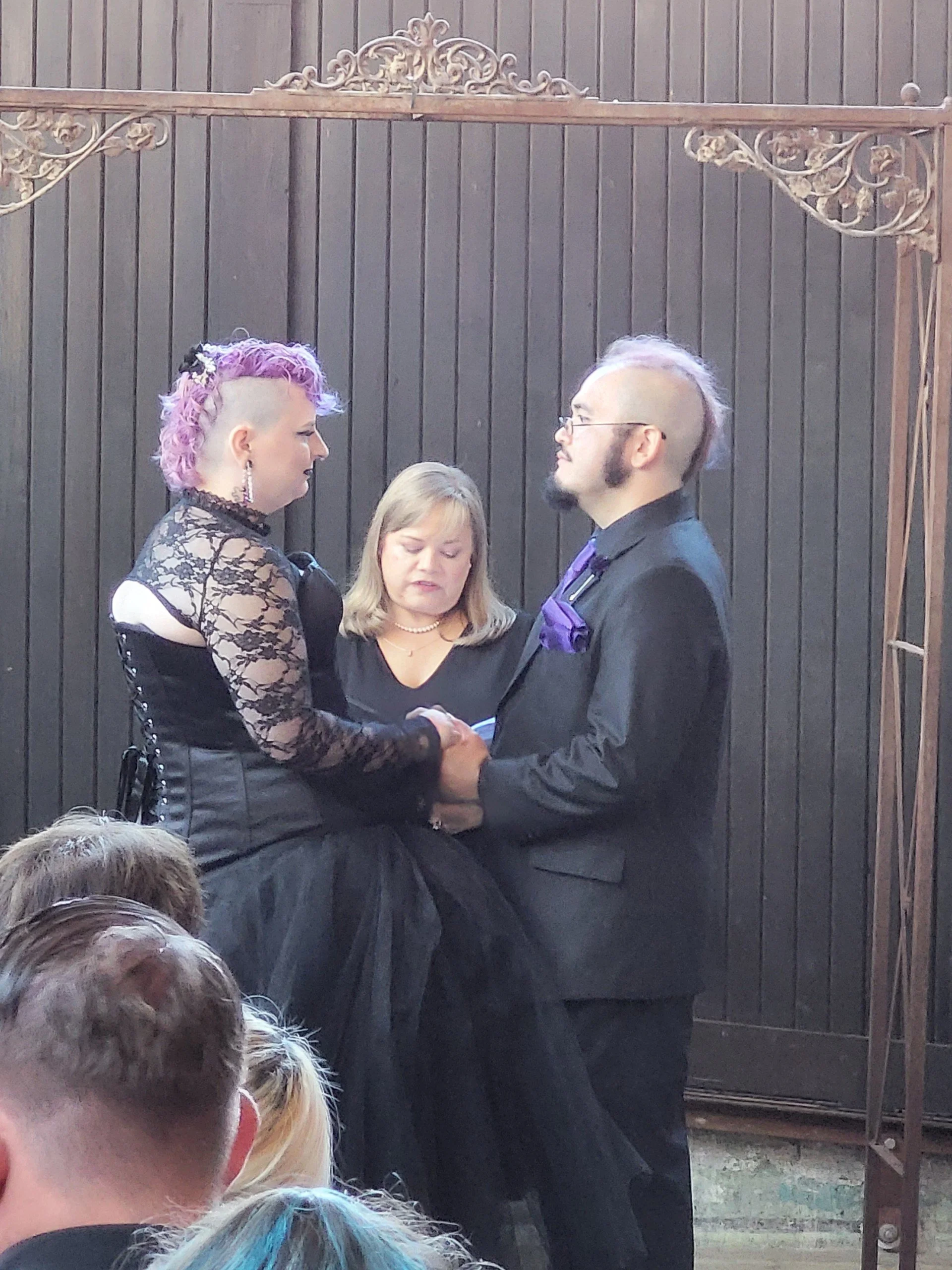 A couple dressed in formal attire, holding hands and facing each other at a wedding ceremony. The woman has pink and purple hair styled in a mohawk, wearing a black lace dress, and the man has a shaved head with a streak of purple hair, wearing glasses, a black suit, and a purple tie with a matching pocket square. They are standing in front of a dark wooden wall with an ornate metal frame above them, likely an outdoor or semi-outdoor setting.