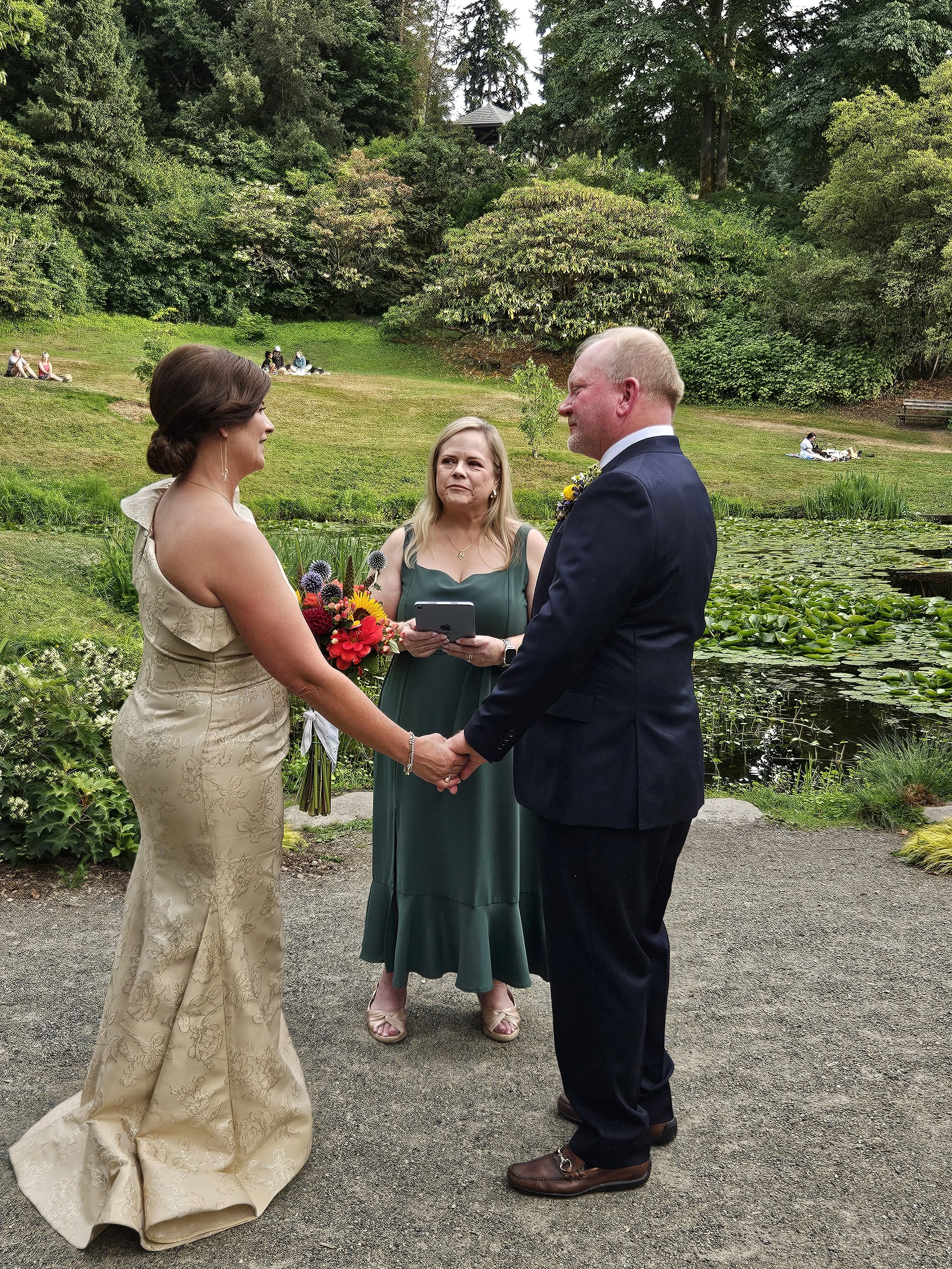 A couple getting married outdoors on a nature trail, holding hands, with an officiant standing between them. The bride is in a light-colored dress, and the groom in a dark suit. They are surrounded by lush greenery and a pond with lily pads.