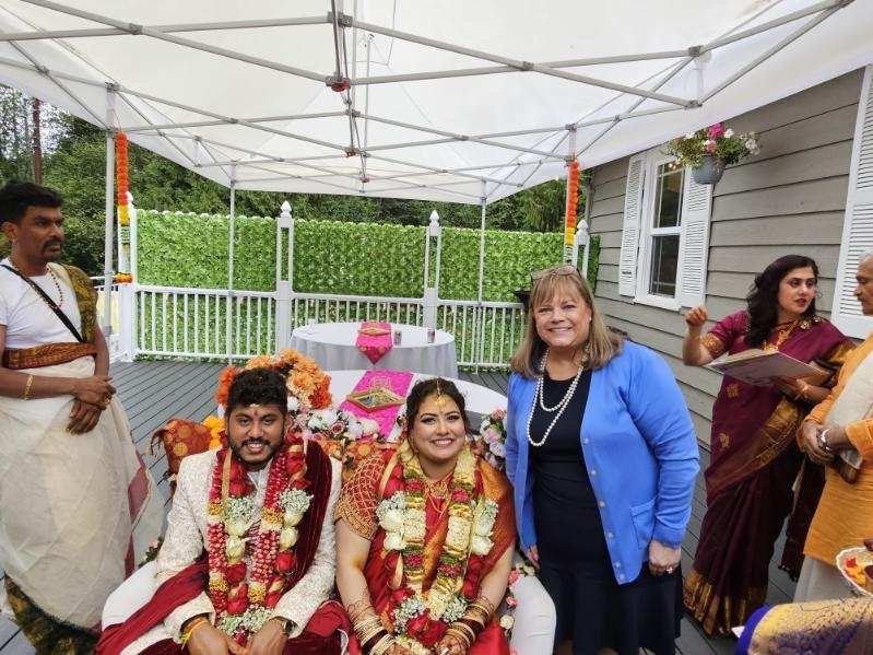 A couple dressed in traditional Indian wedding attire seated under a white canopy with floral decorations, surrounded by guests. A woman in a blue blazer stands next to the couple smiling. The setting appears to be an outdoor event.