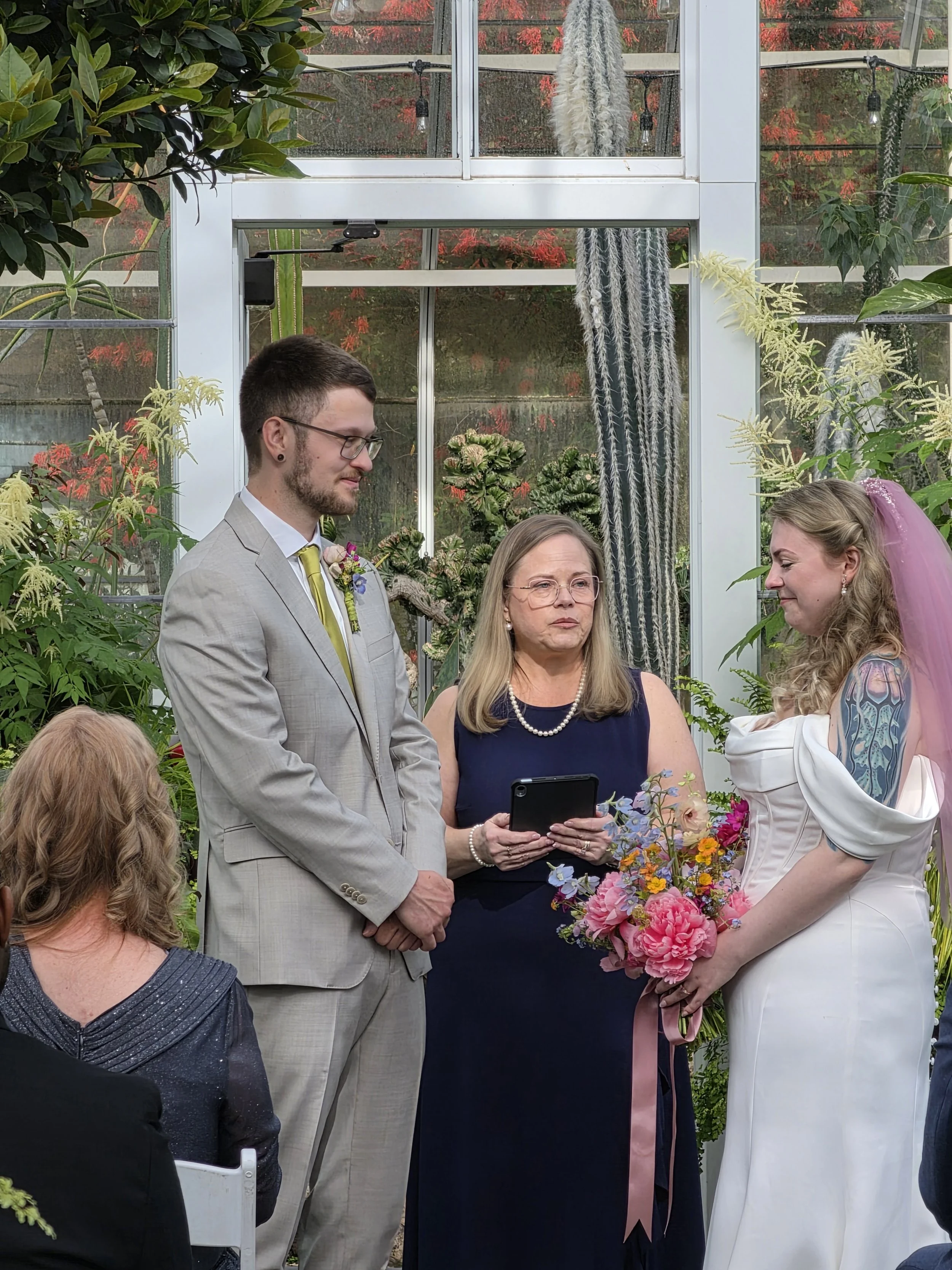 A wedding ceremony in a greenhouse with a groom in a light gray suit and glasses, and a bride in a white dress with a pink veil, holding a bouquet of pink and purple flowers. An officiant in a dark dress is between them, holding a tablet. Guests are 