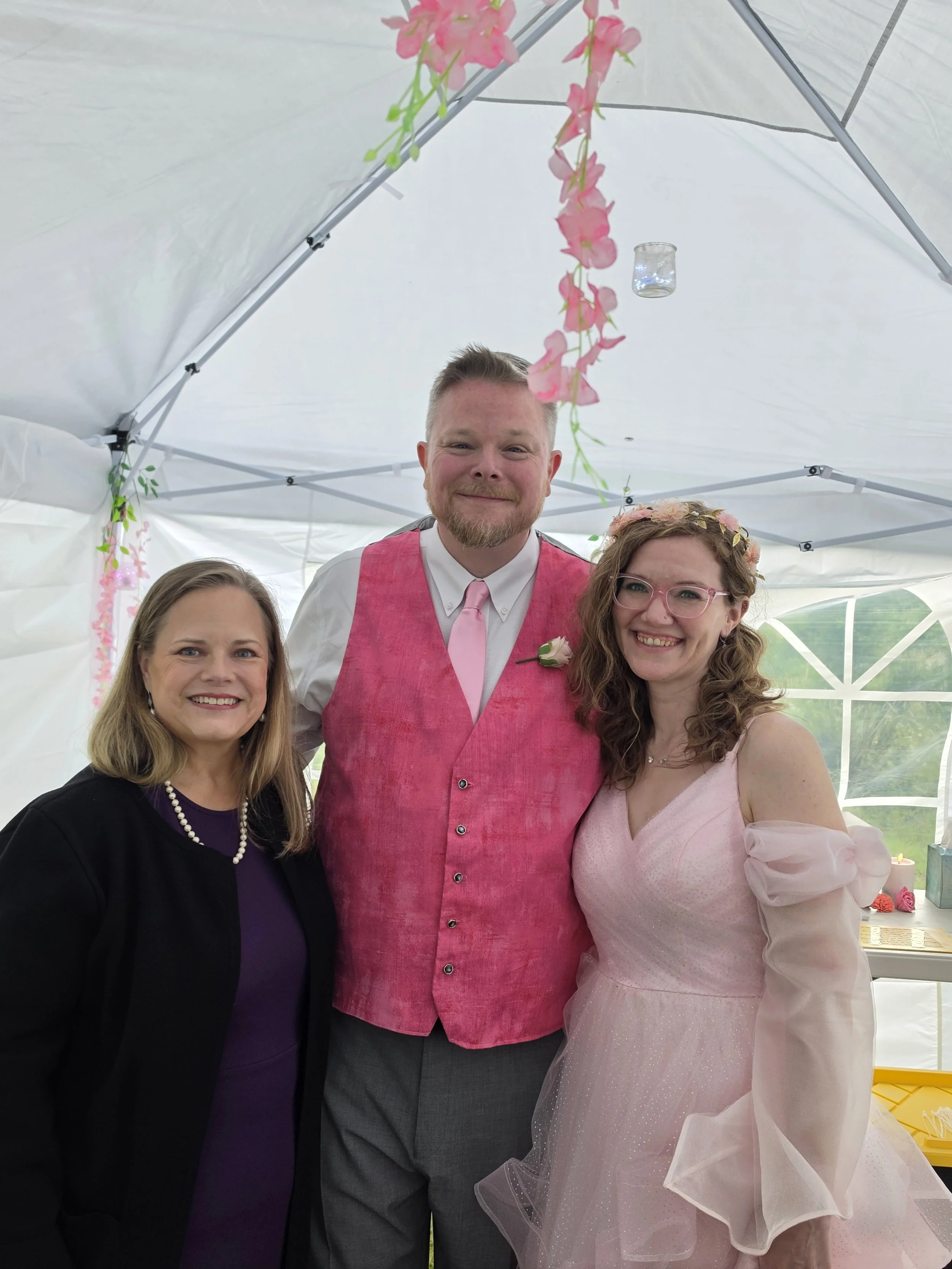 A group of four people posing together inside a white canopy tent. There are two women on either side and a man in the middle. The man is wearing a pink vest and pink tie, with a white shirt, and has short blonde hair and a beard. The women are smili