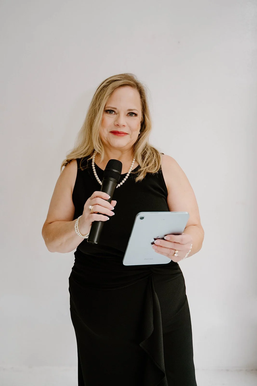 A woman with blonde hair wearing a black dress with pearl jewelry holding a microphone and a tablet, standing against a plain white background.