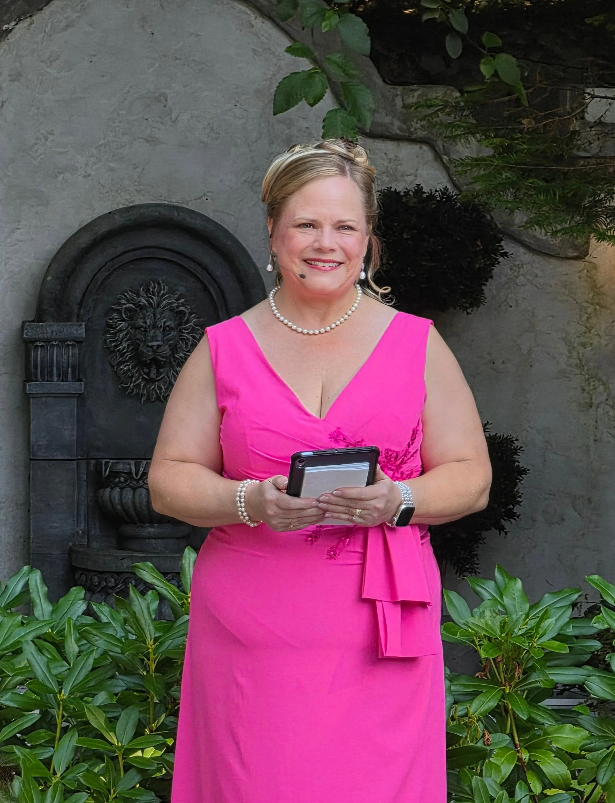 A woman in a bright pink dress with pearl jewelry, holding a smartphone, standing outdoors in front of greenery and a decorative wall fountain.