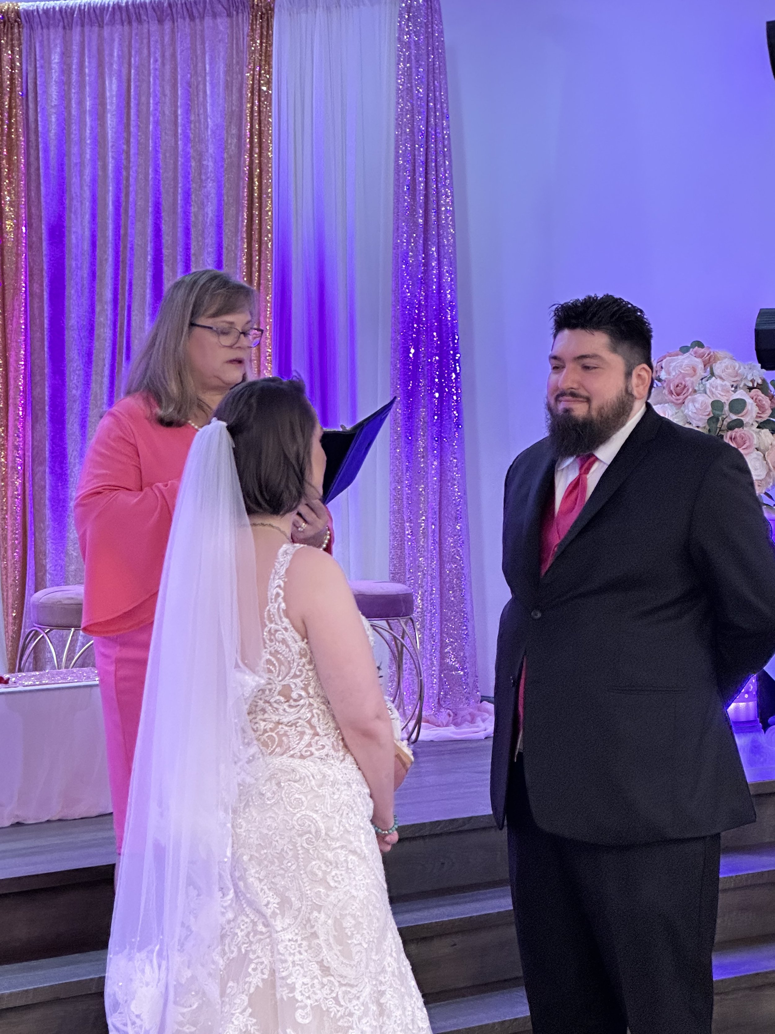 A wedding ceremony with a bride in a white dress facing a groom in a black suit with a red tie, officiated by a woman in a pink top, standing in front of a decorated backdrop.