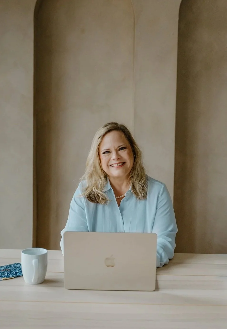 A woman with blonde hair smiling at a laptop on a wooden desk, with a white coffee mug and a smartphone with a blue blue case nearby.
