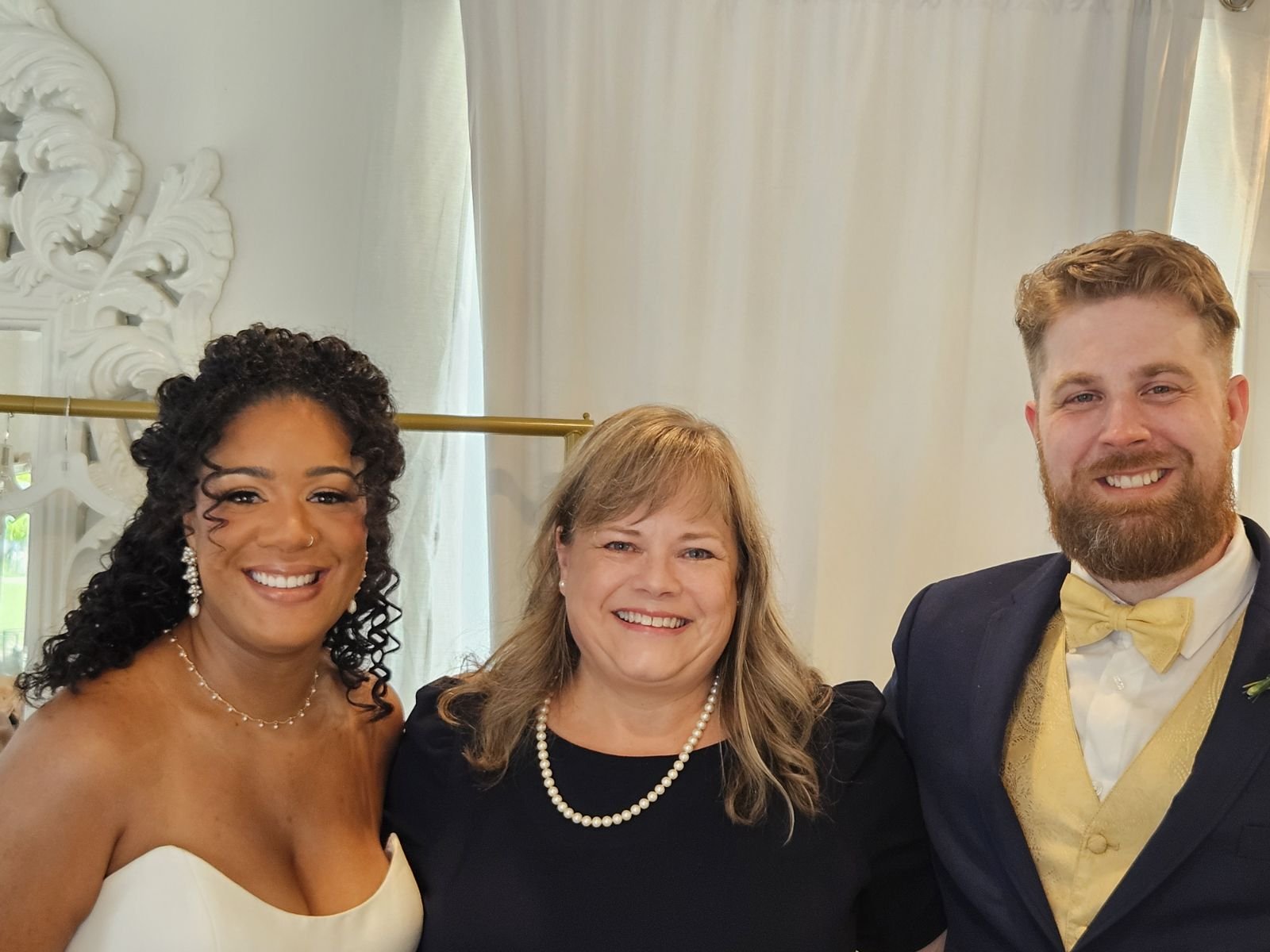 Three people smiling at a formal event; a woman in a white dress, a woman in a dark dress, and a man in a suit with a yellow vest and bow tie.