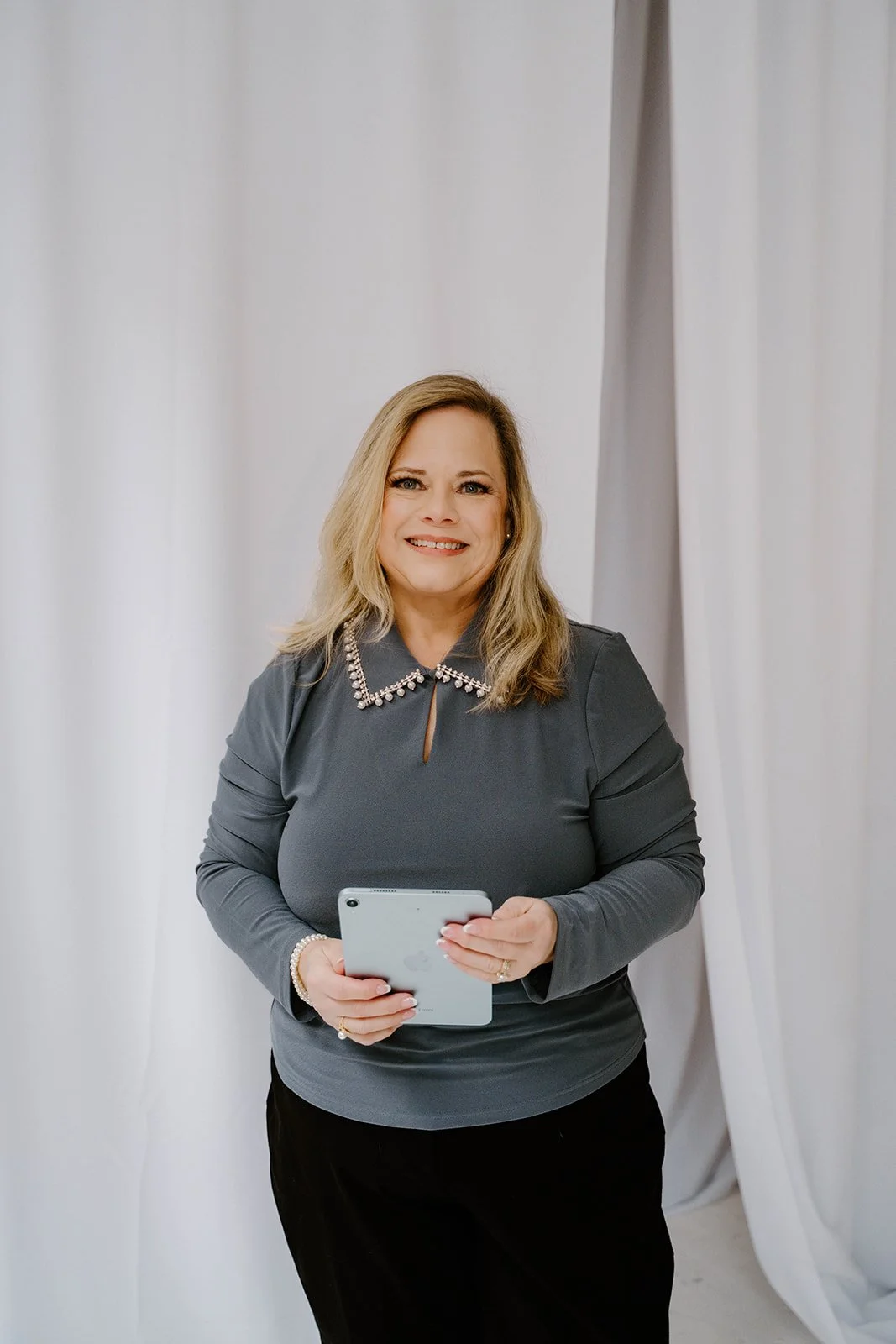Woman with blonde hair smiling, holding a tablet, wearing a dark gray top with a pearl-studded collar, standing in front of white curtains.