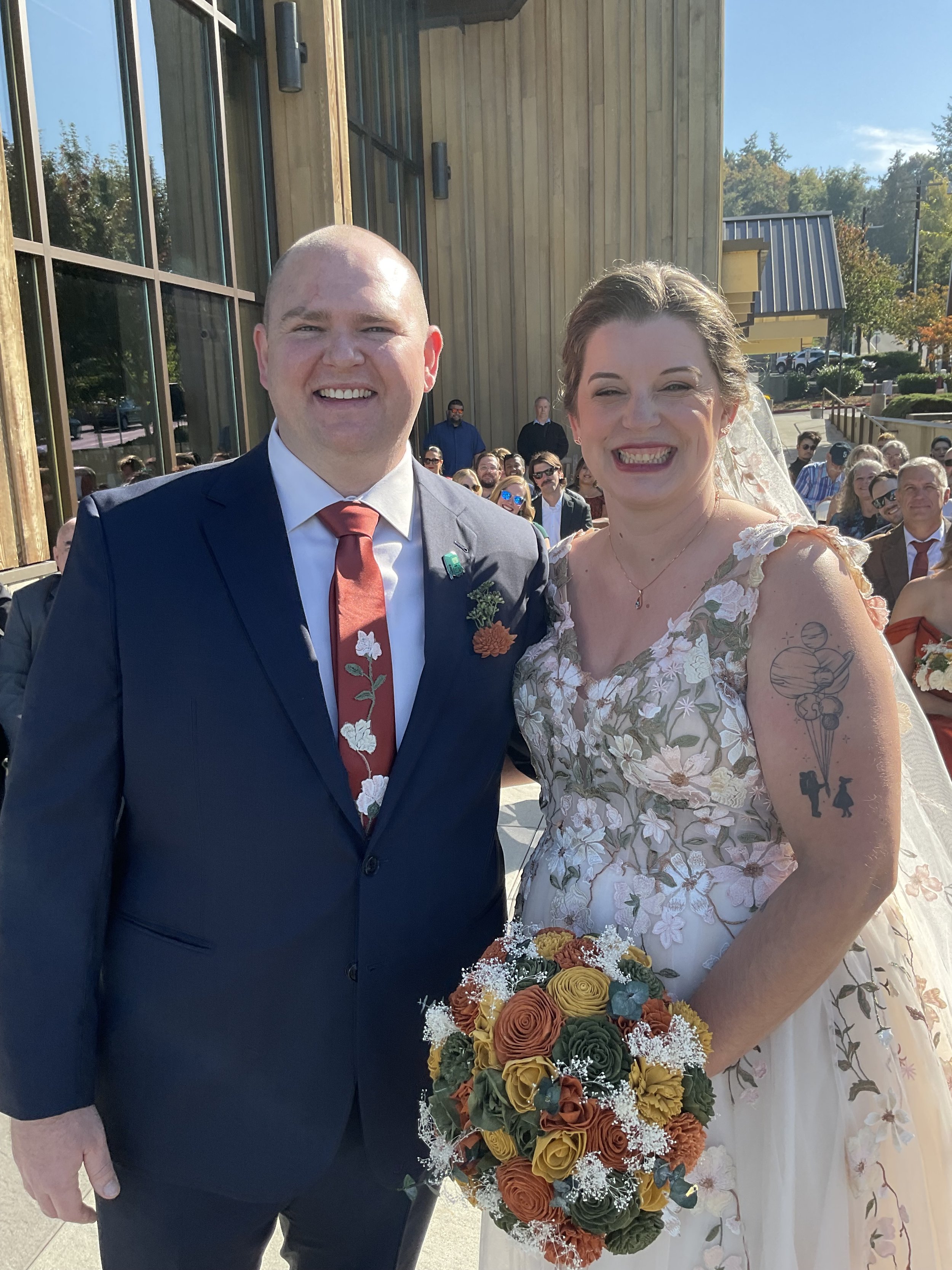 A smiling couple at an outdoor wedding, the groom is wearing a dark suit with a floral tie, and the bride is in a floral gown holding a bouquet of colorful flowers. Guests are seated in the background.