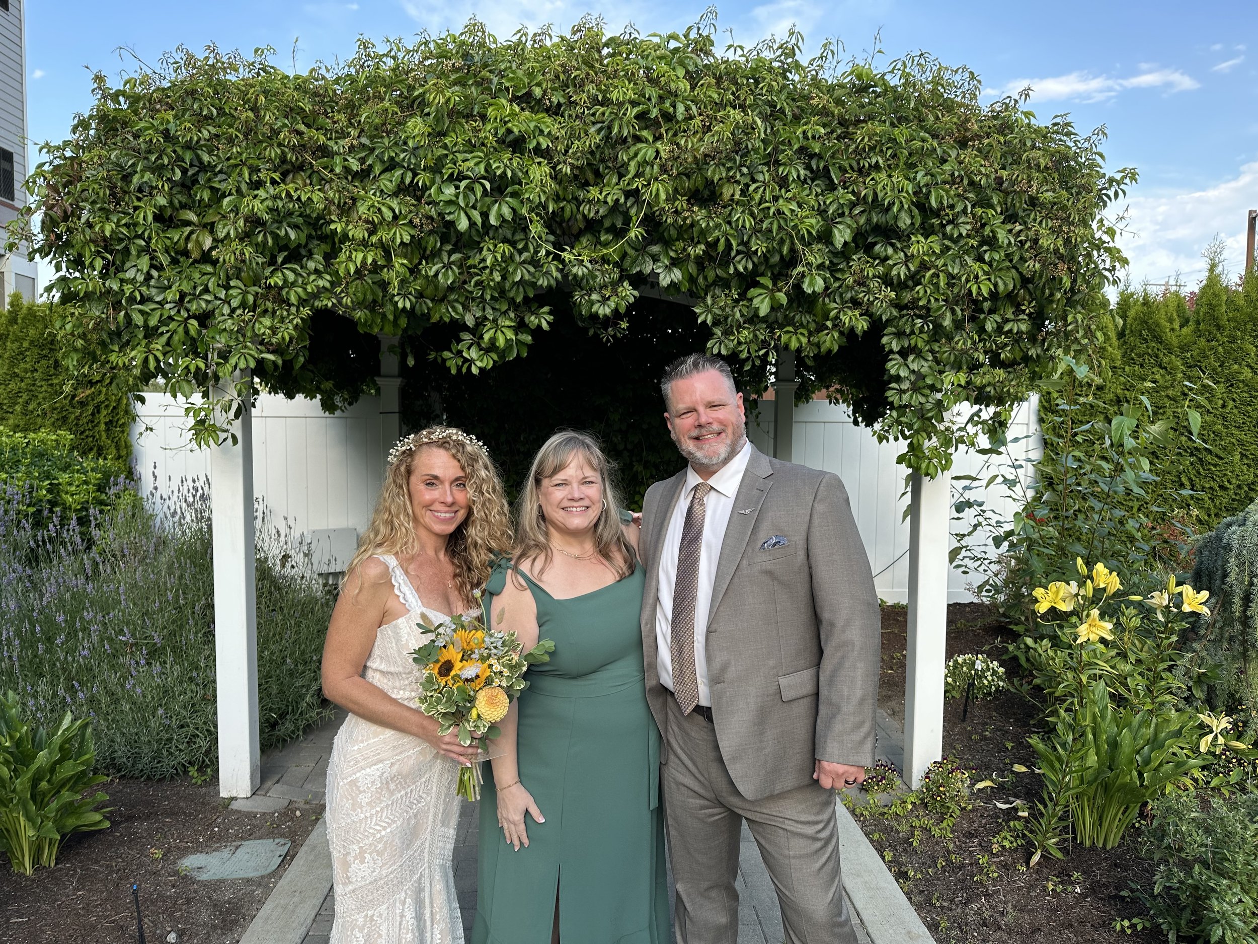A bride in a white dress holding a bouquet stands beside a woman in a green dress and a man in a suit under a greenery-covered pergola.