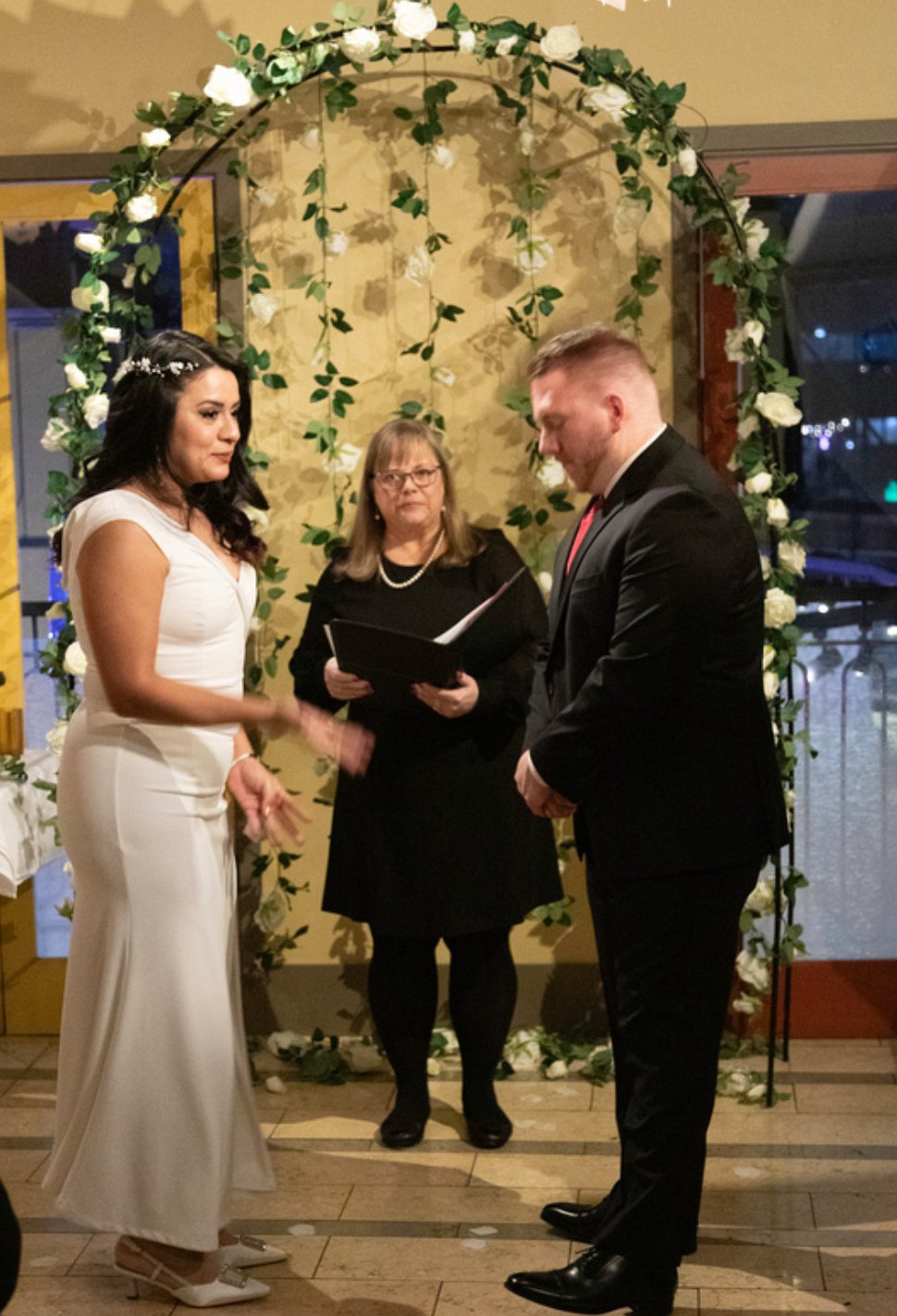 A wedding ceremony with a bride in a white dress and a groom in a black suit standing under a floral arch, with an officiant in black holding a book.