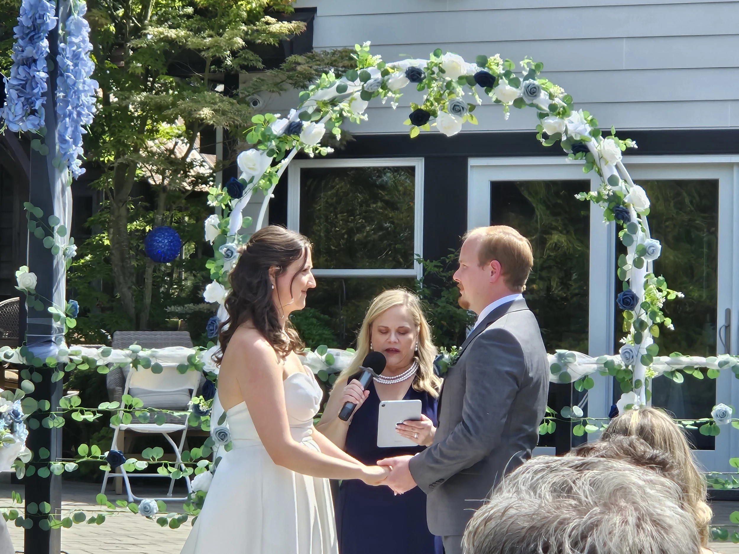 A couple getting married during an outdoor wedding ceremony, standing under a floral arch with an officiant and guests in the background.