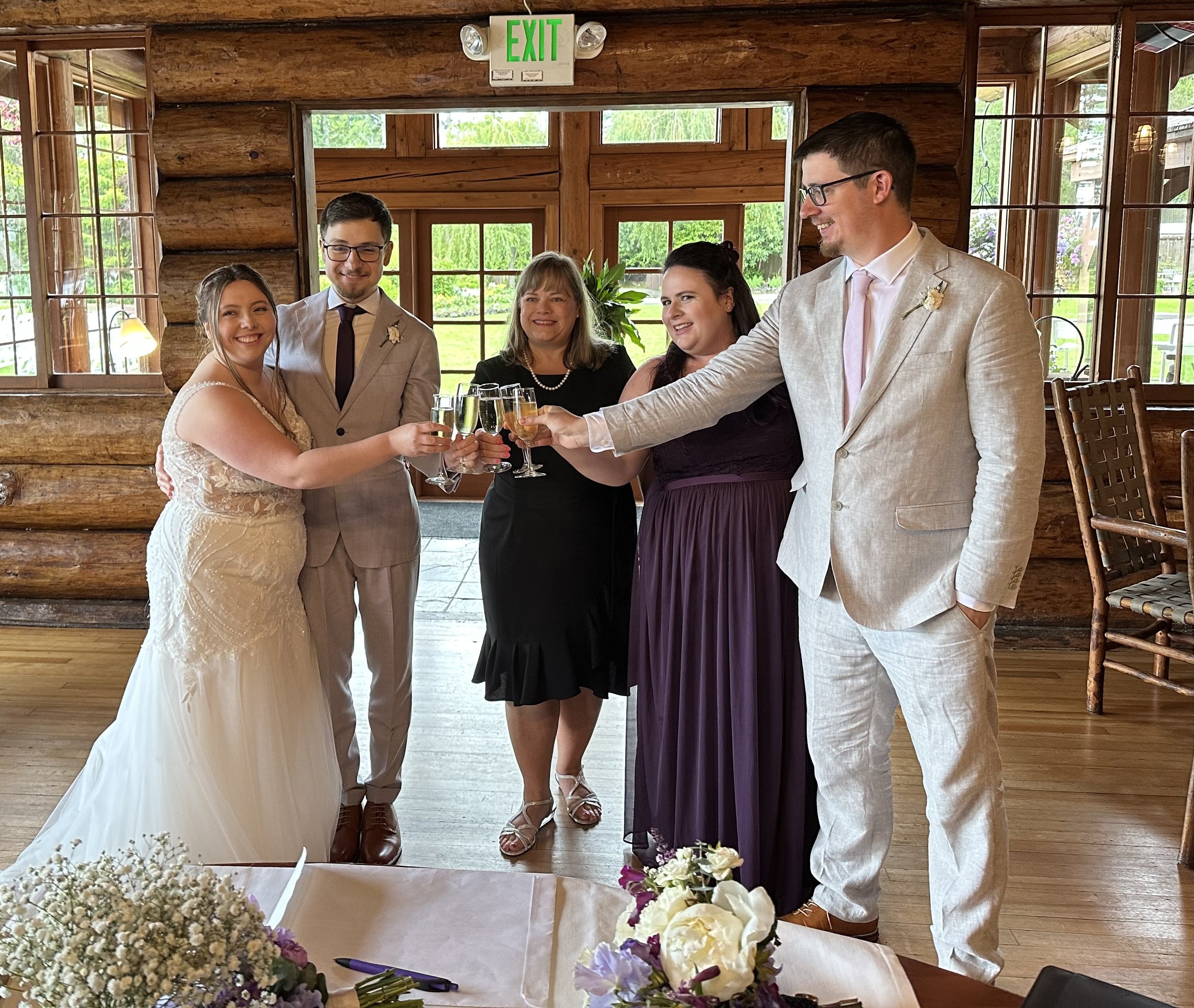 Group of people toasting at a wedding, with the bride in a white dress and other guests dressed formally, standing in a rustic venue.