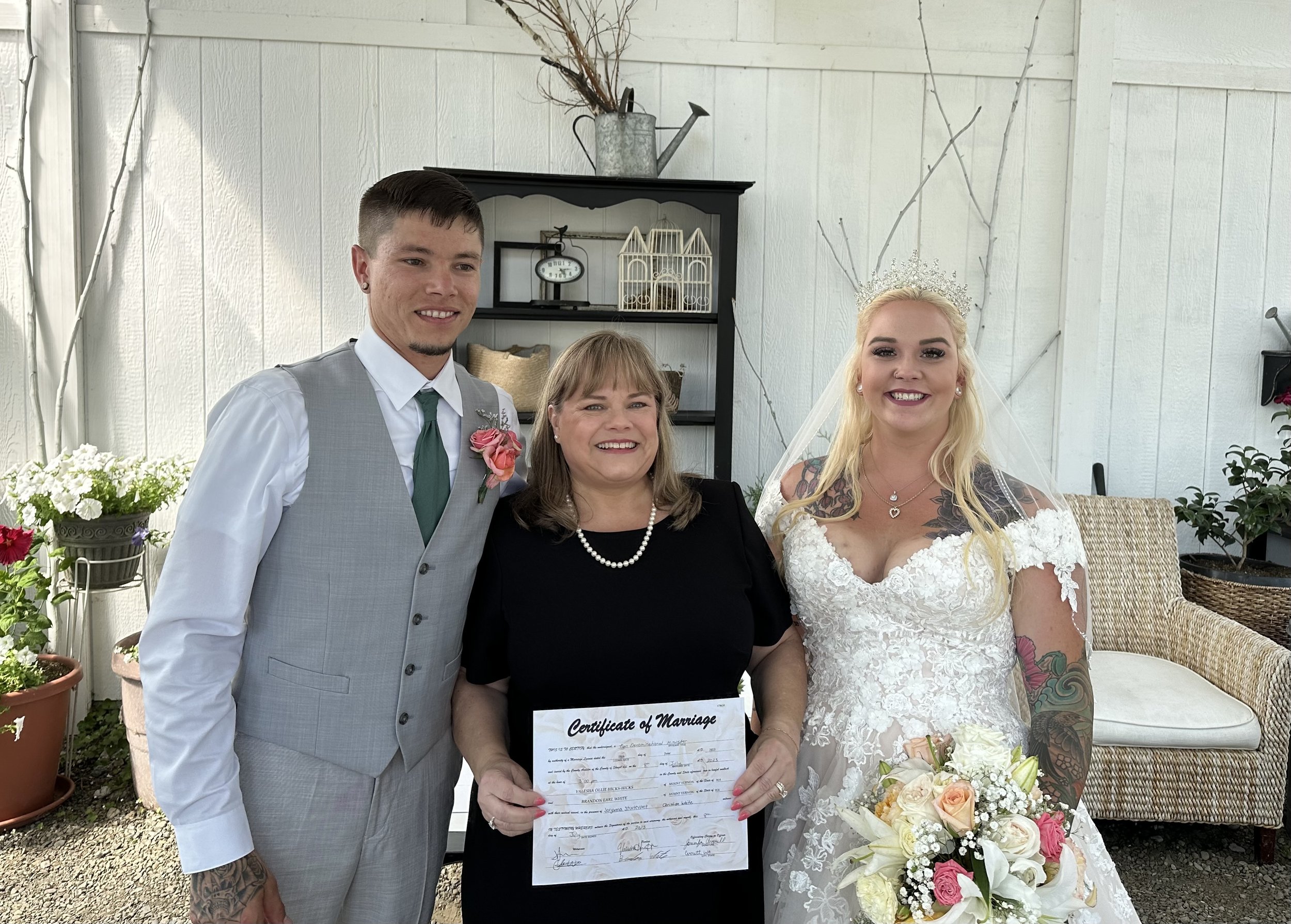 A wedding couple posing with an officiant holding a certificate of marriage, standing in a decorated indoor venue. The groom is in a gray suit, the bride in a white lace gown, holding a bouquet of flowers.