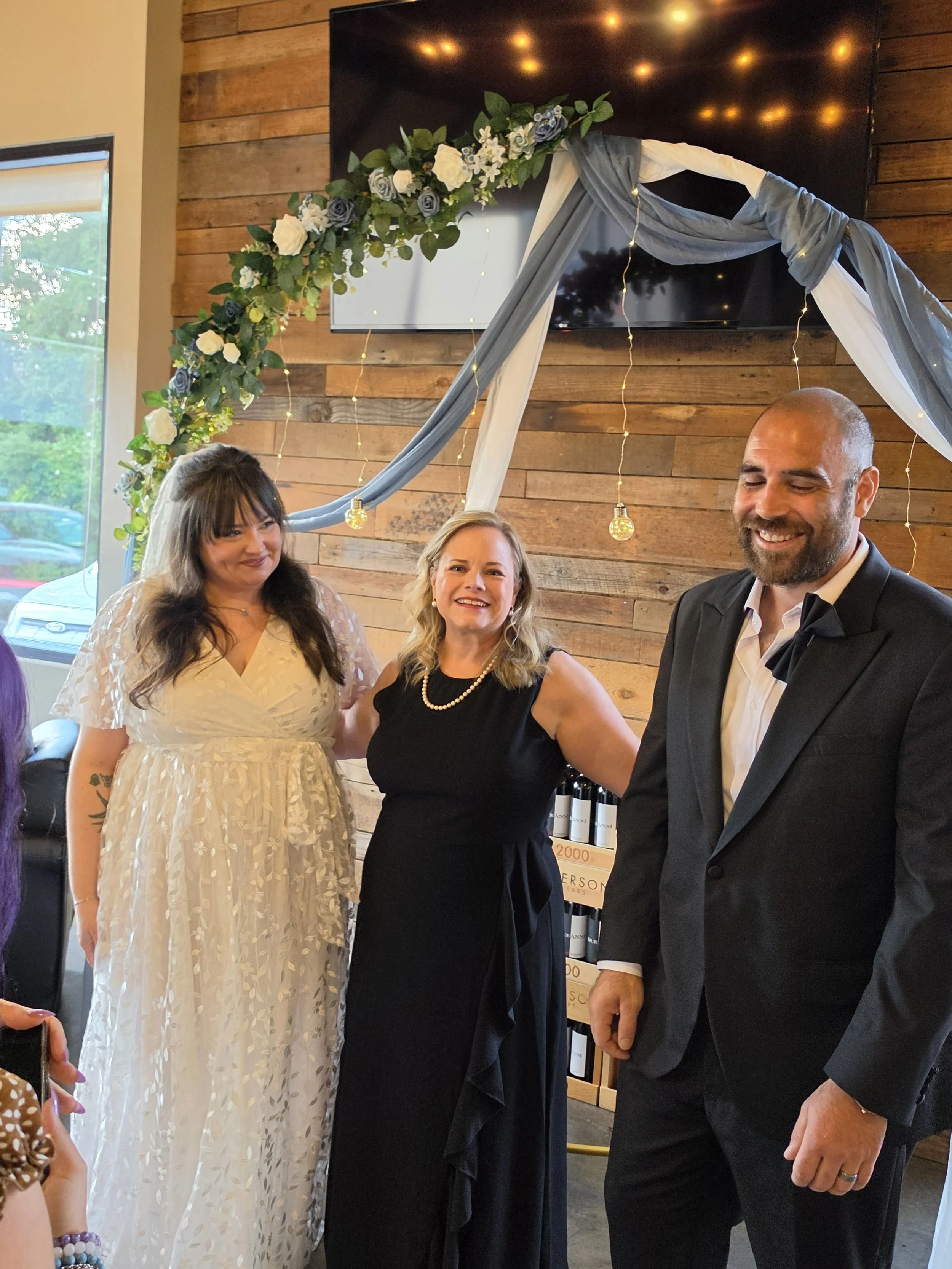 People standing together at an event, with a decorated floral arch above them, in a room with wood panel walls and large TV screen behind them, during daytime.