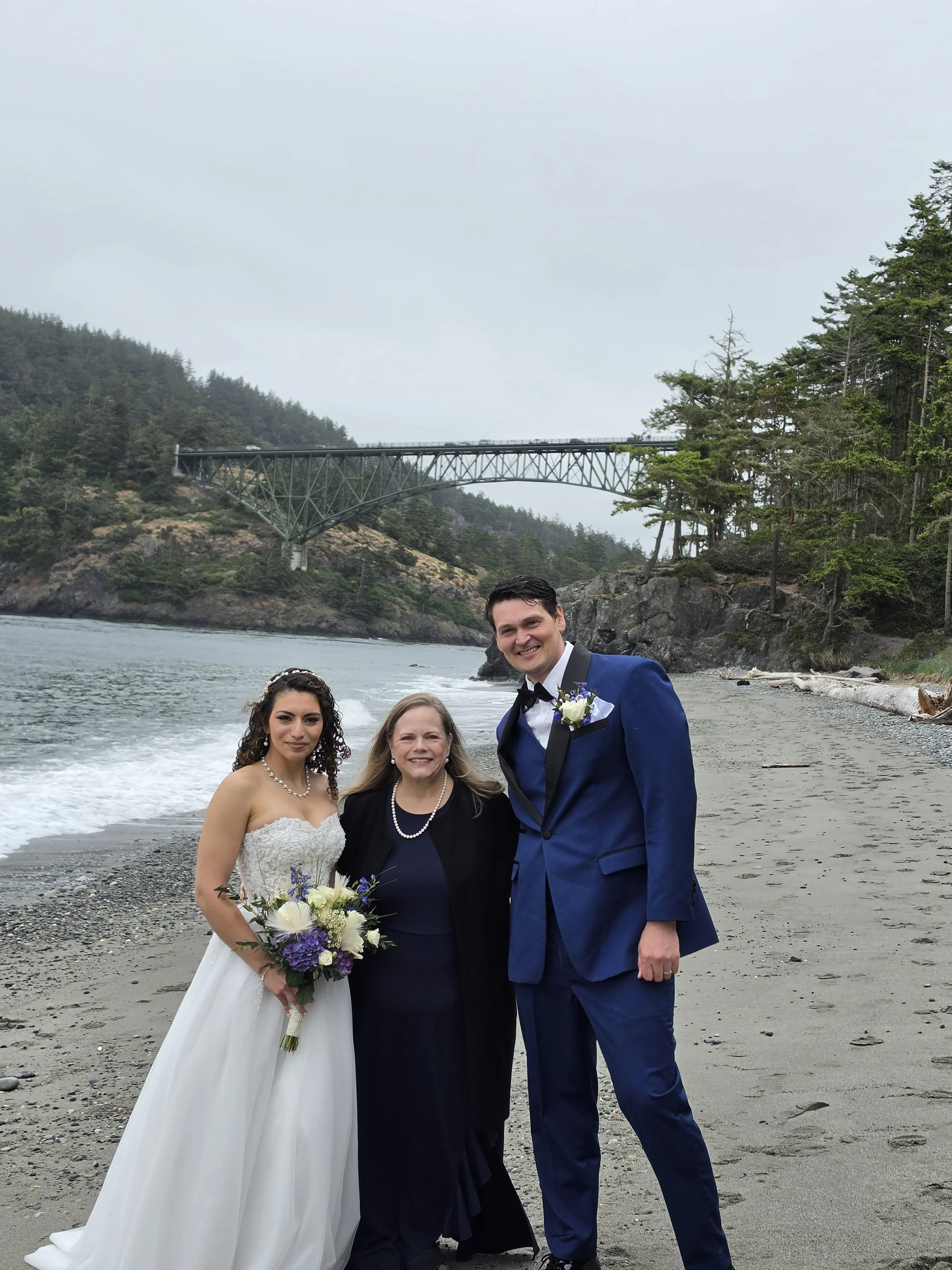 A bride, groom, and two women standing on a beach. The bride is in a white wedding gown holding a bouquet, and the groom is in a blue suit with a boutonniere. One woman is in a black dress and the other is in a black blazer. Behind them is a rocky shoreline, trees, and a bridge over the water.