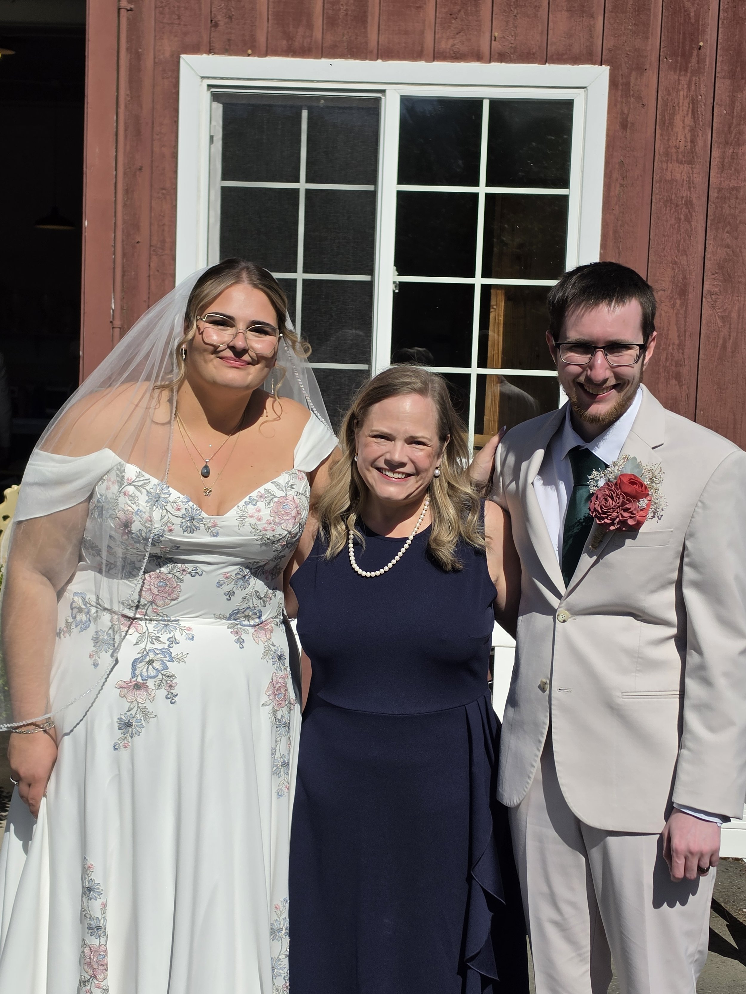 Three people posing outdoors in front of a red wooden building with a window behind them. The woman on the left is wearing a white dress with floral embroidery, a veil, and glasses. The woman in the middle is wearing a dark blue dress and pearl neckl