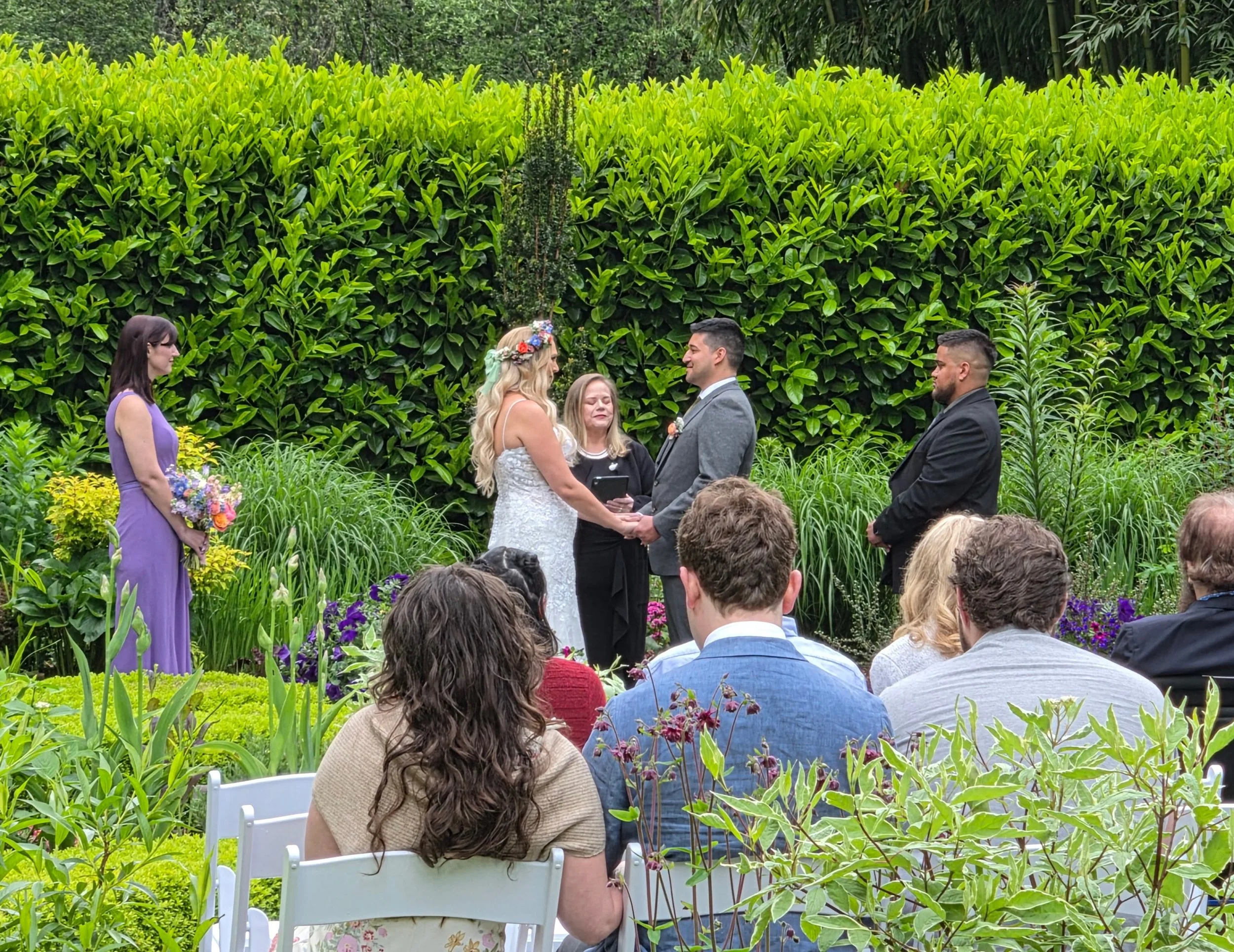 A wedding ceremony taking place outdoors in a garden, with a bride and groom holding hands and exchanging vows. The bride is wearing a white dress with a floral crown, and the groom is in a gray suit. An officiant and a bridesmaid in a purple dress h