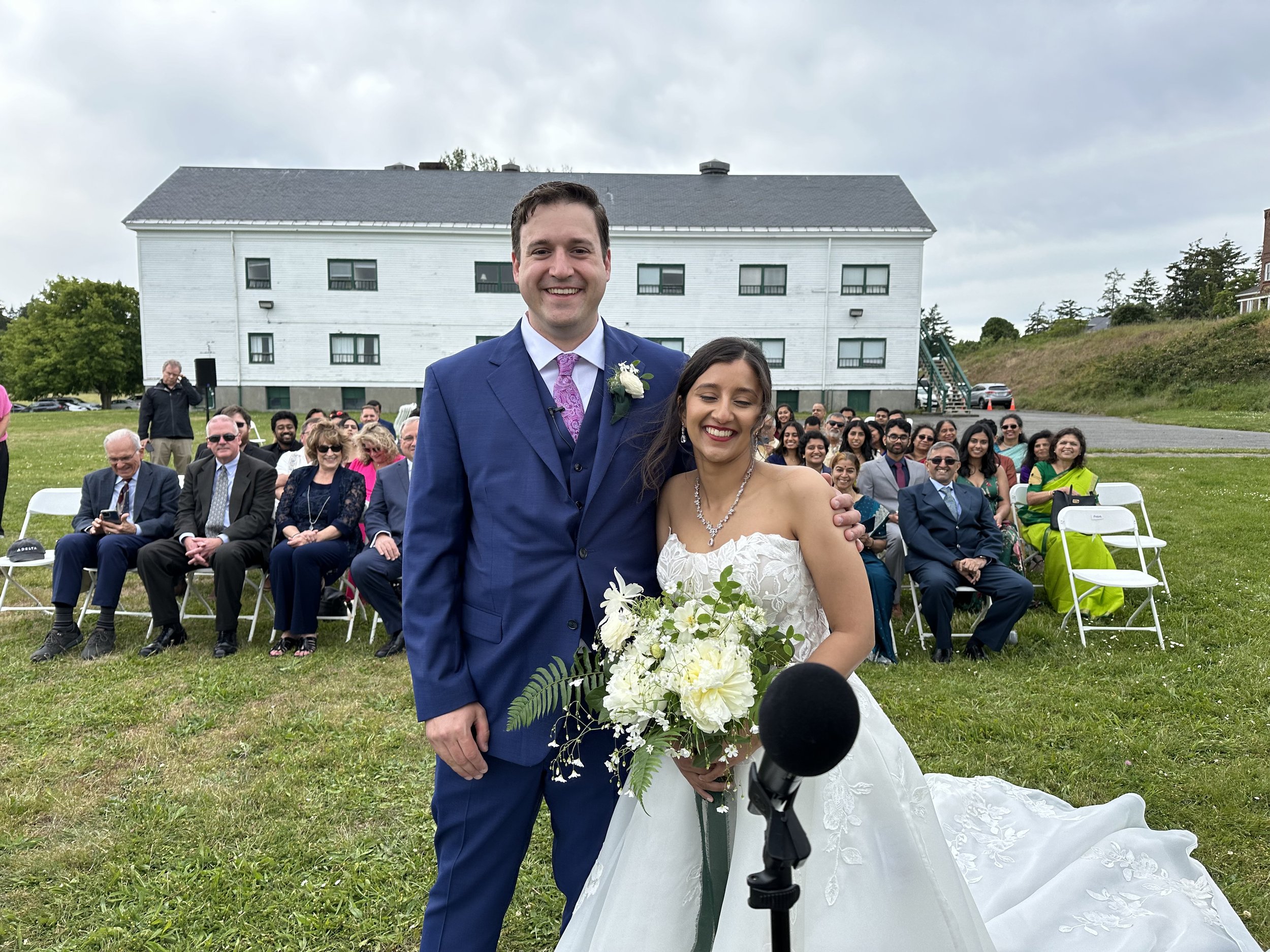 A smiling bride and groom standing outdoors with a bouquet, surrounded by guests seated in rows on a grassy area. A white building is visible in the background.