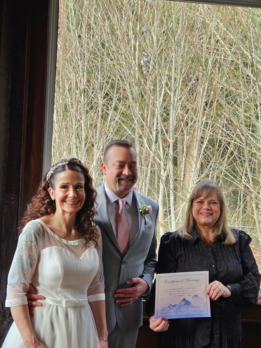 A bride in a white dress standing next to a groom in a gray suit with a pink tie. They are smiling and posing with a woman in black holding a marriage certificate. The background shows leafless trees outside a window.