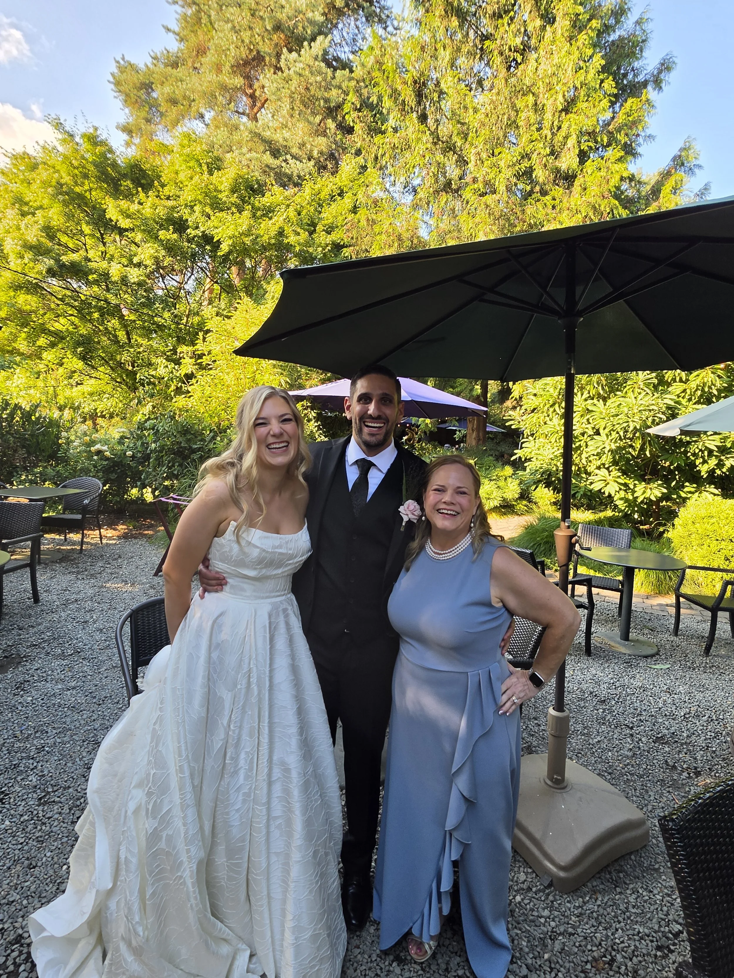 Three people smiling and standing outdoors under a black umbrella, with lush green trees and a blue sky in the background. The woman on the left is in a white dress, the man in the center is in a black suit, and the woman on the right is in a light b