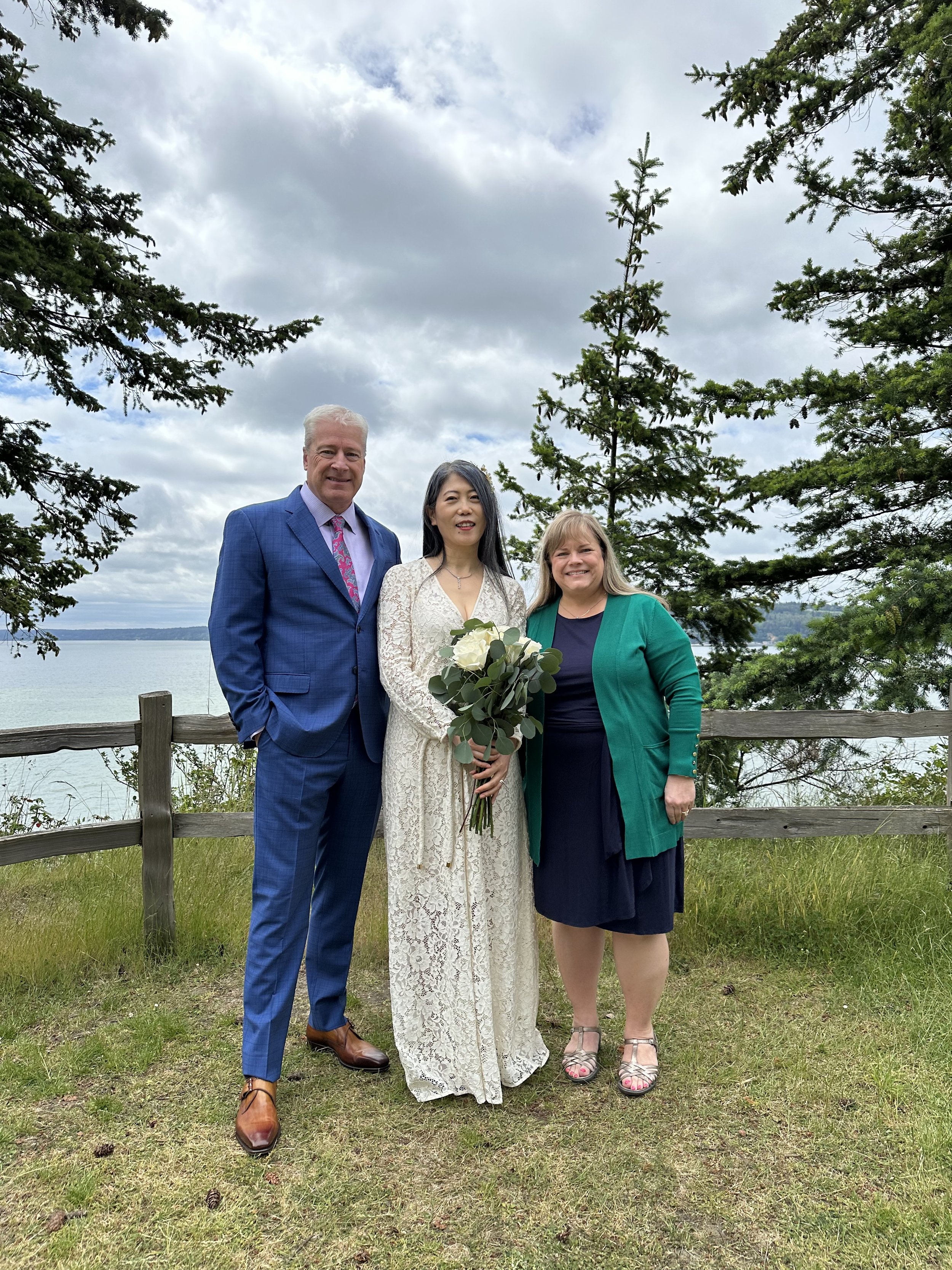 Three people, including a bride in a lace dress holding a bouquet, stand outdoors near a fence with pine trees and a body of water in the background.