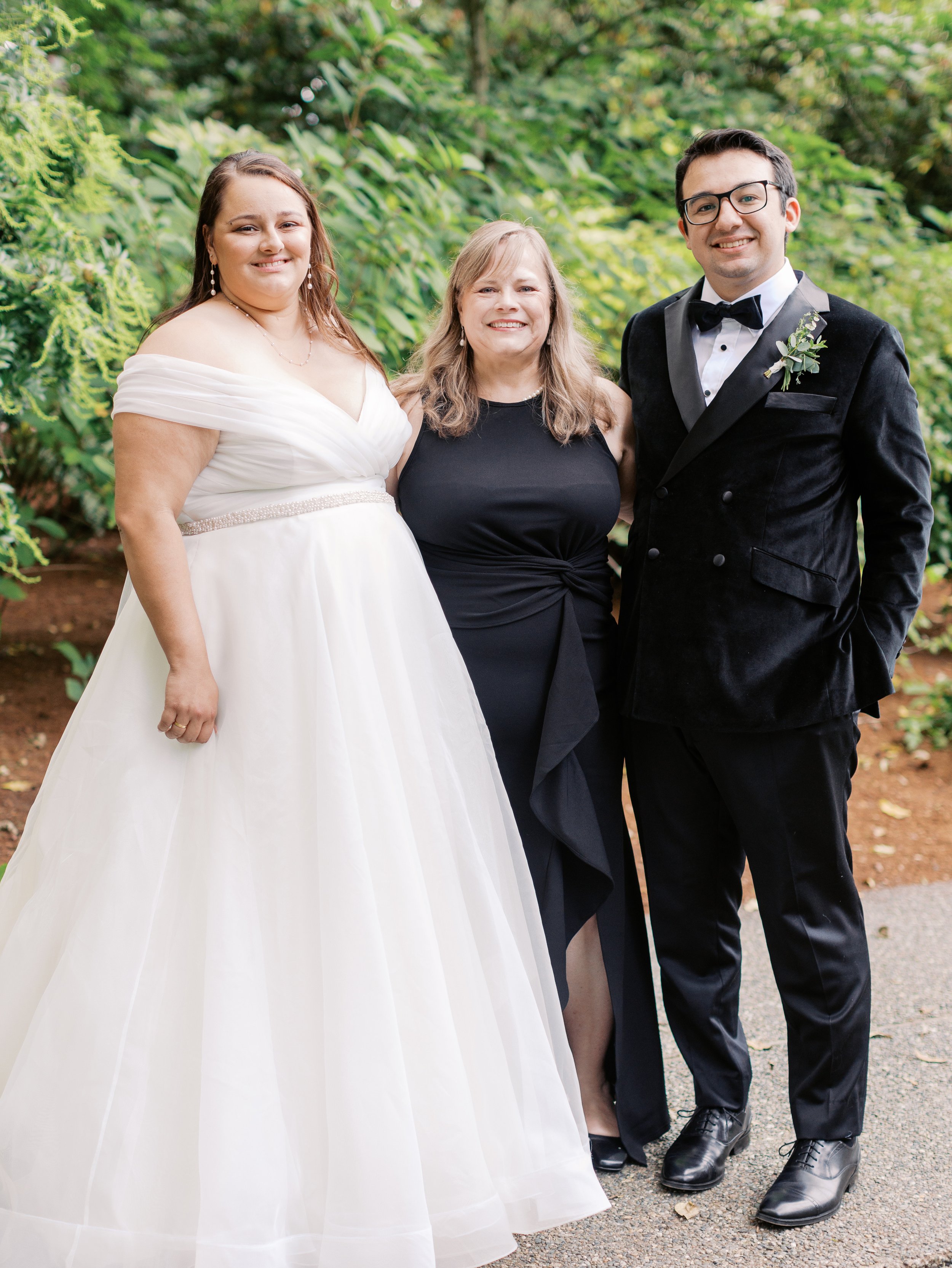 Three people posing outdoors; a bride in a white wedding dress, a woman in a black dress, and a man in a black tuxedo.