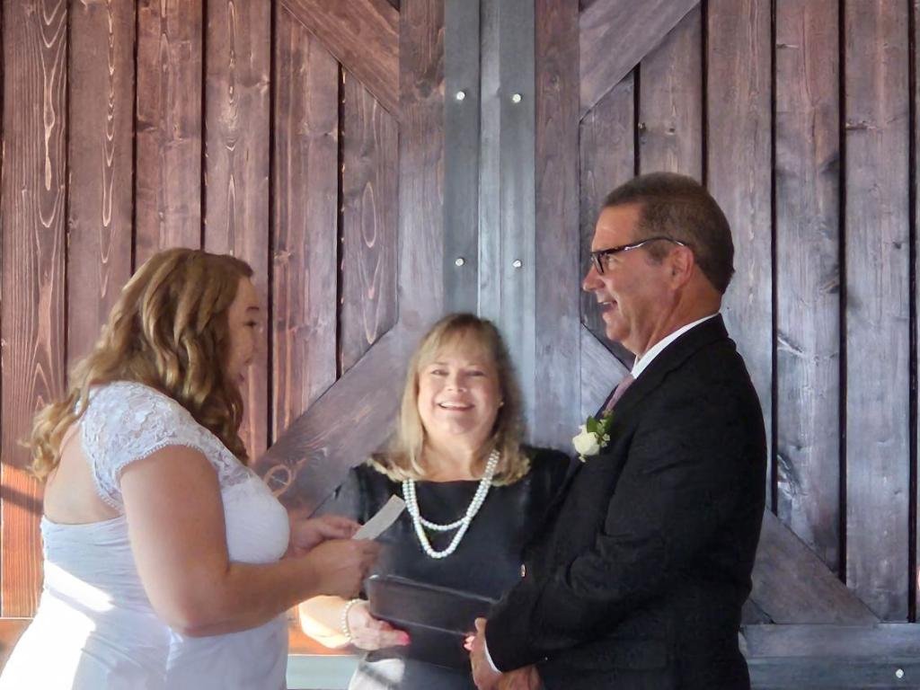 A wedding ceremony with a bride and groom exchanging vows in front of a wooden barn door. The bride is wearing a white dress, and the groom is in a suit. An officiant stands between them, smiling.