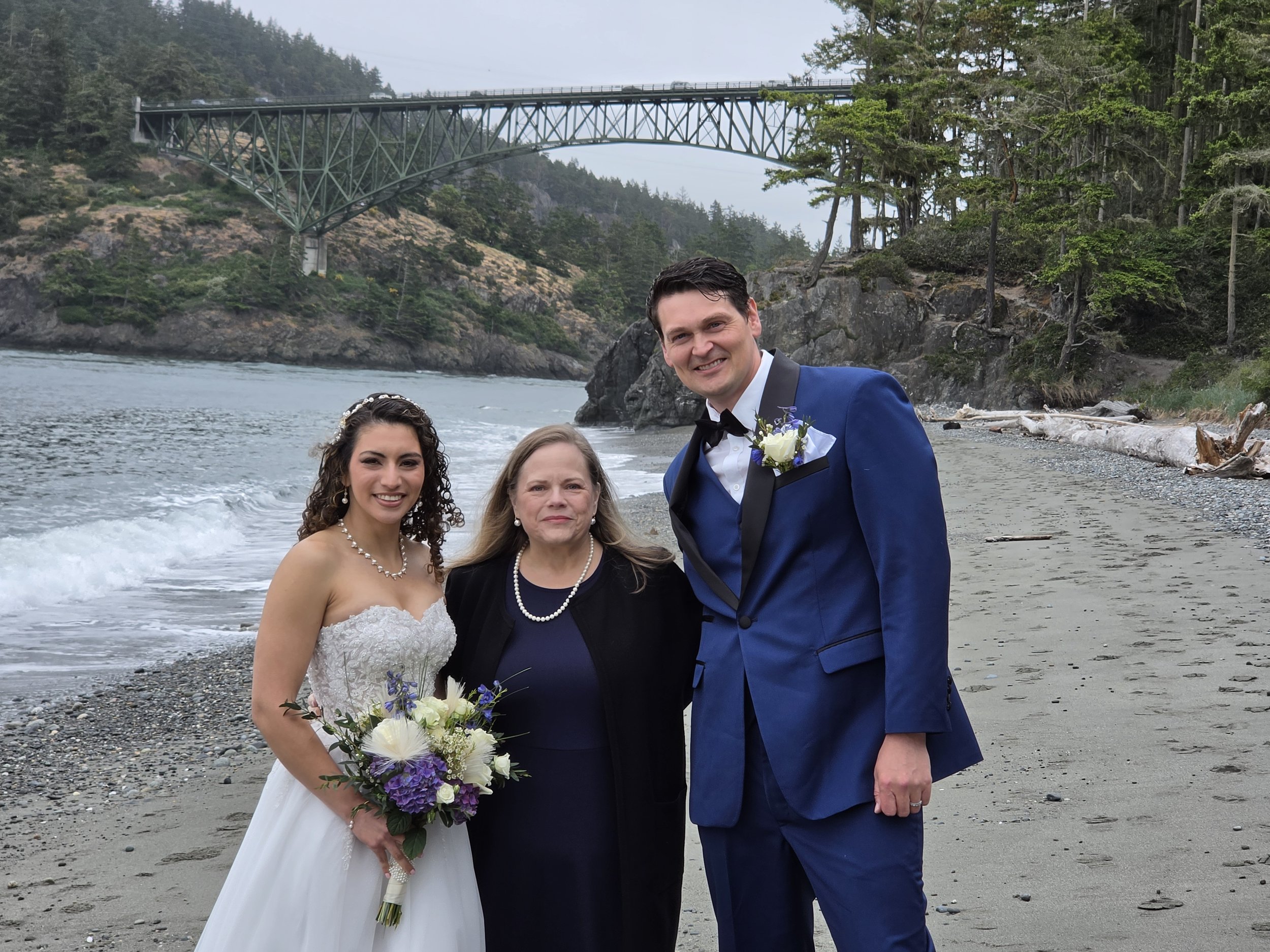 A bride and groom at the beach, with two women, one in black and another in white, standing in front of them. The groom is wearing a blue tuxedo with a black bow tie, and the bride is in a strapless wedding gown holding a bouquet of white and purple 