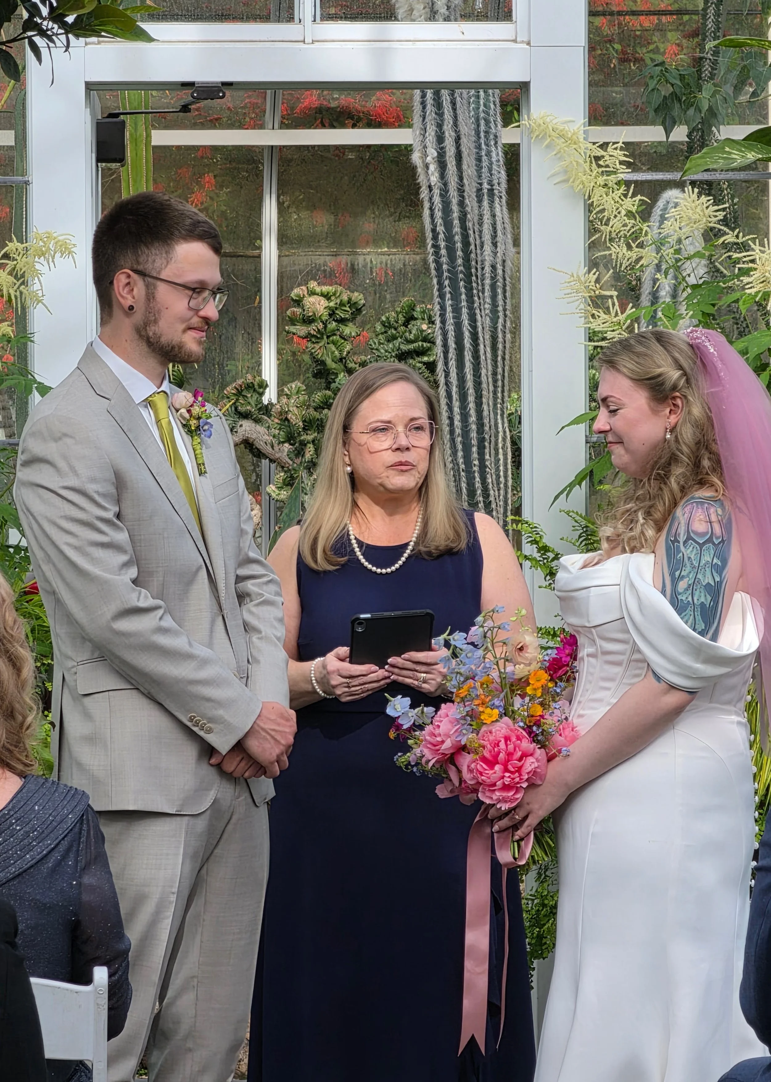 A wedding ceremony in a greenhouse with a groom, a bride, and an officiant. The groom is dressed in a light tan suit with a yellow tie and a boutonniere. The bride is wearing a white wedding dress with a pink veil and holding a bouquet of pink and purple flowers. The officiant, a woman, is standing between them holding a phone, and they are all surrounded by green plants and flowers.