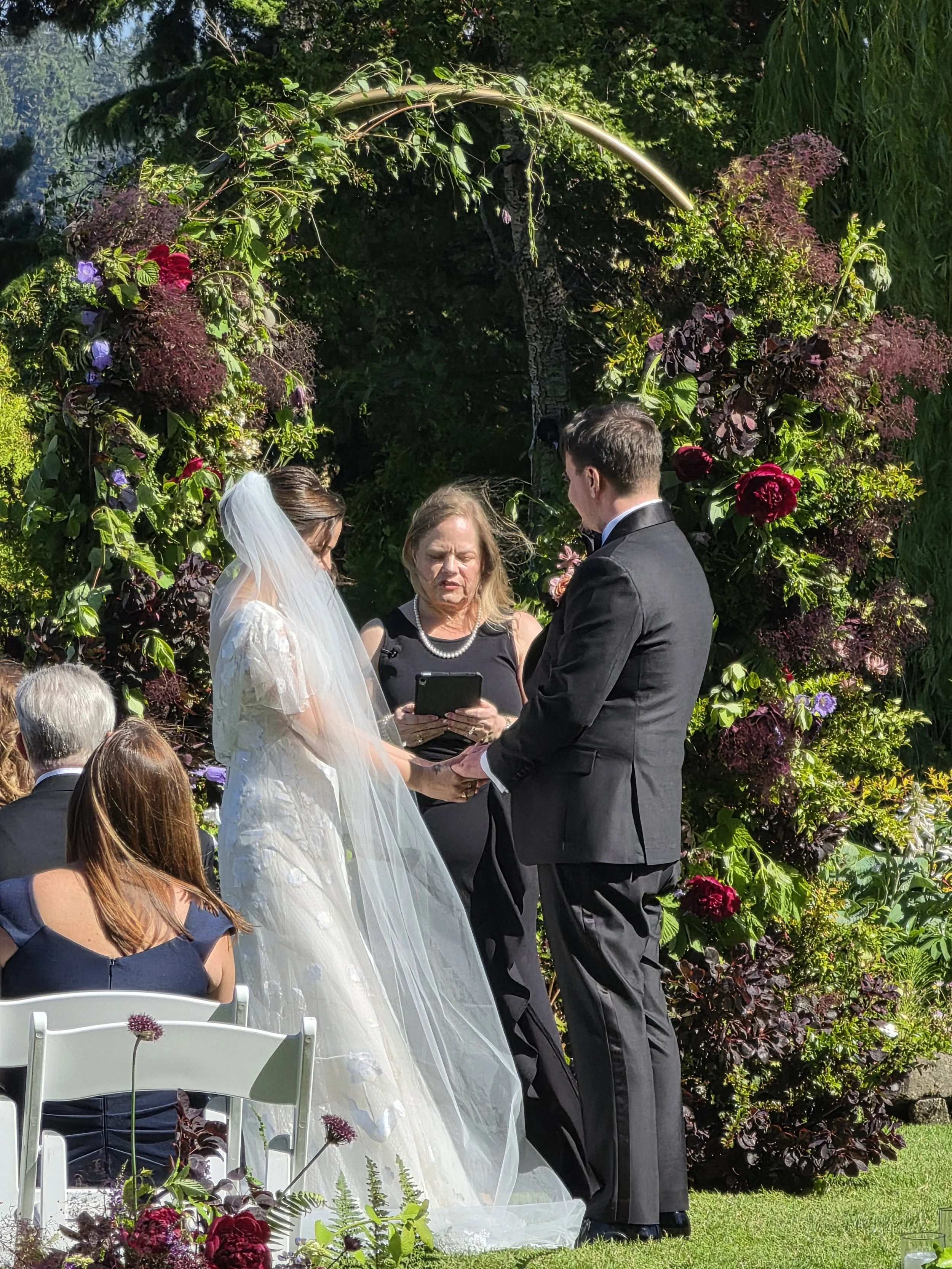 A wedding ceremony outdoors with a bride and groom facing each other, exchanging vows, in front of a floral arch. An officiant stands between them, reading from a tablet. Guests are seated in chairs, and the background features lush greenery and flow