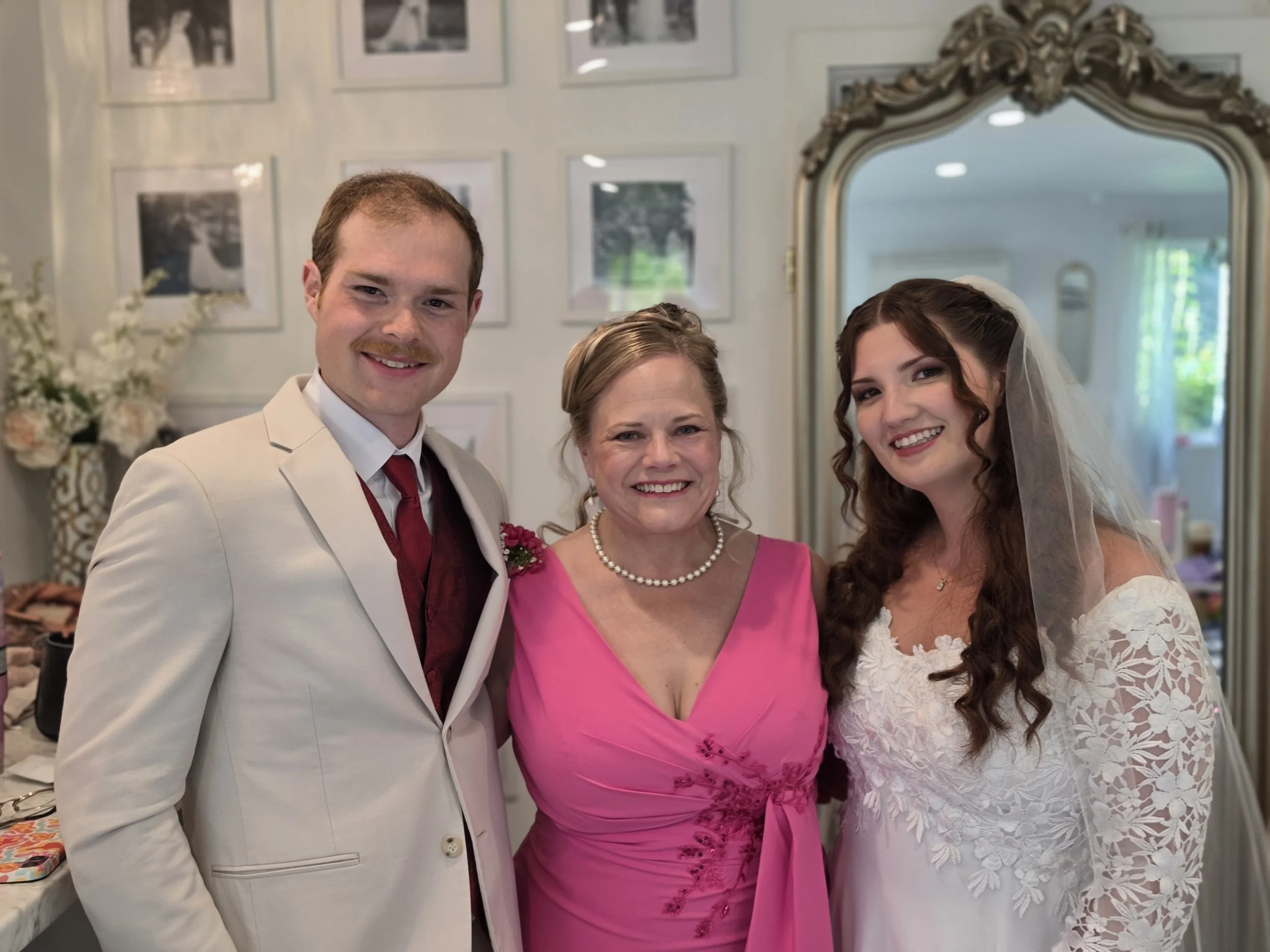 A group of four people smiling for a photo at a wedding celebration. The group includes a man wearing a light-colored suit with a red tie, a woman in a pink dress with pearls, a woman in a white lace wedding dress with a veil, and a man in the backgr