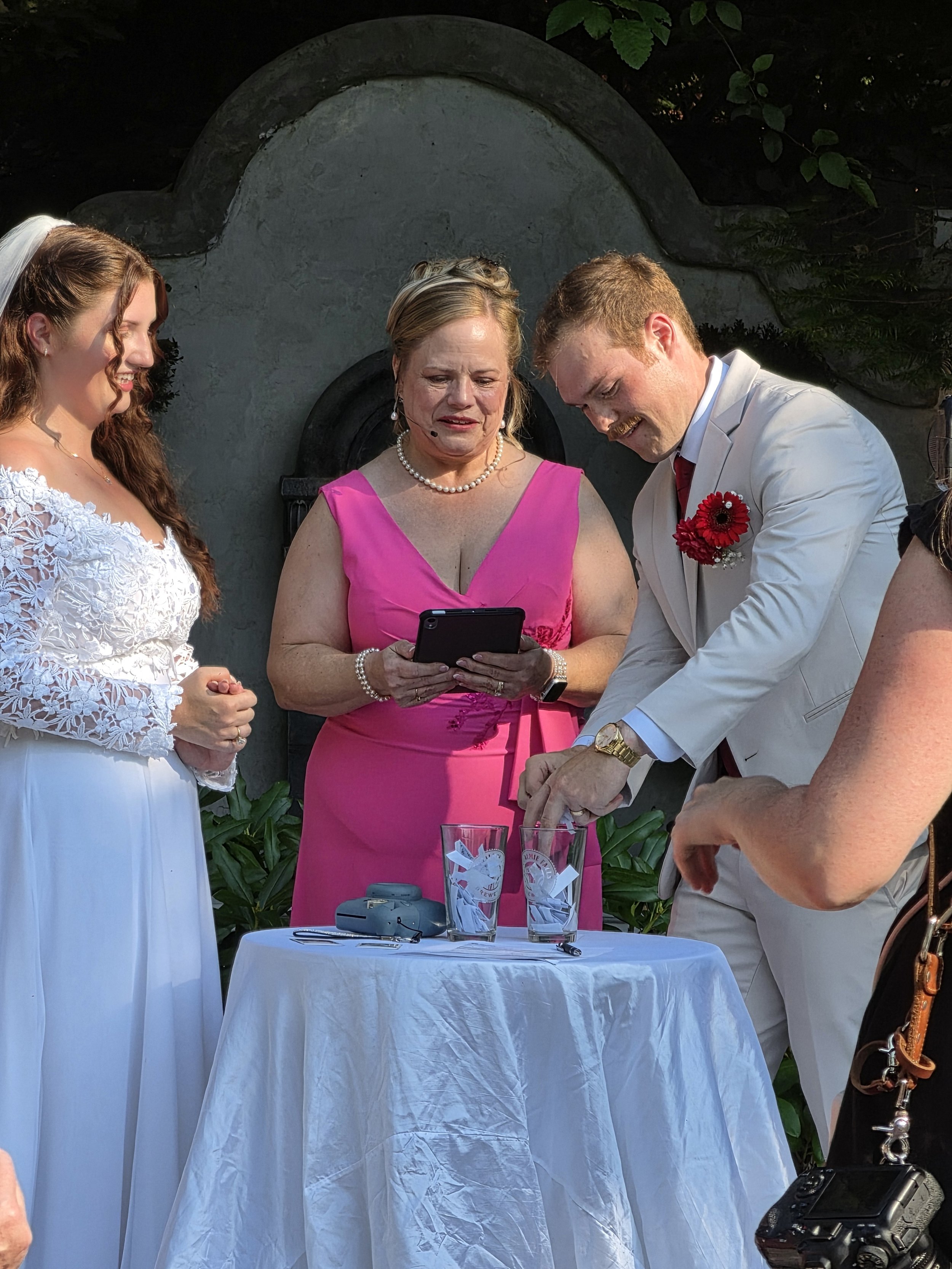 A wedding ceremony with the bride and groom standing at a table, looking at a phone held by a woman in a pink dress, with an officiant in the center, and guests partially visible around them.