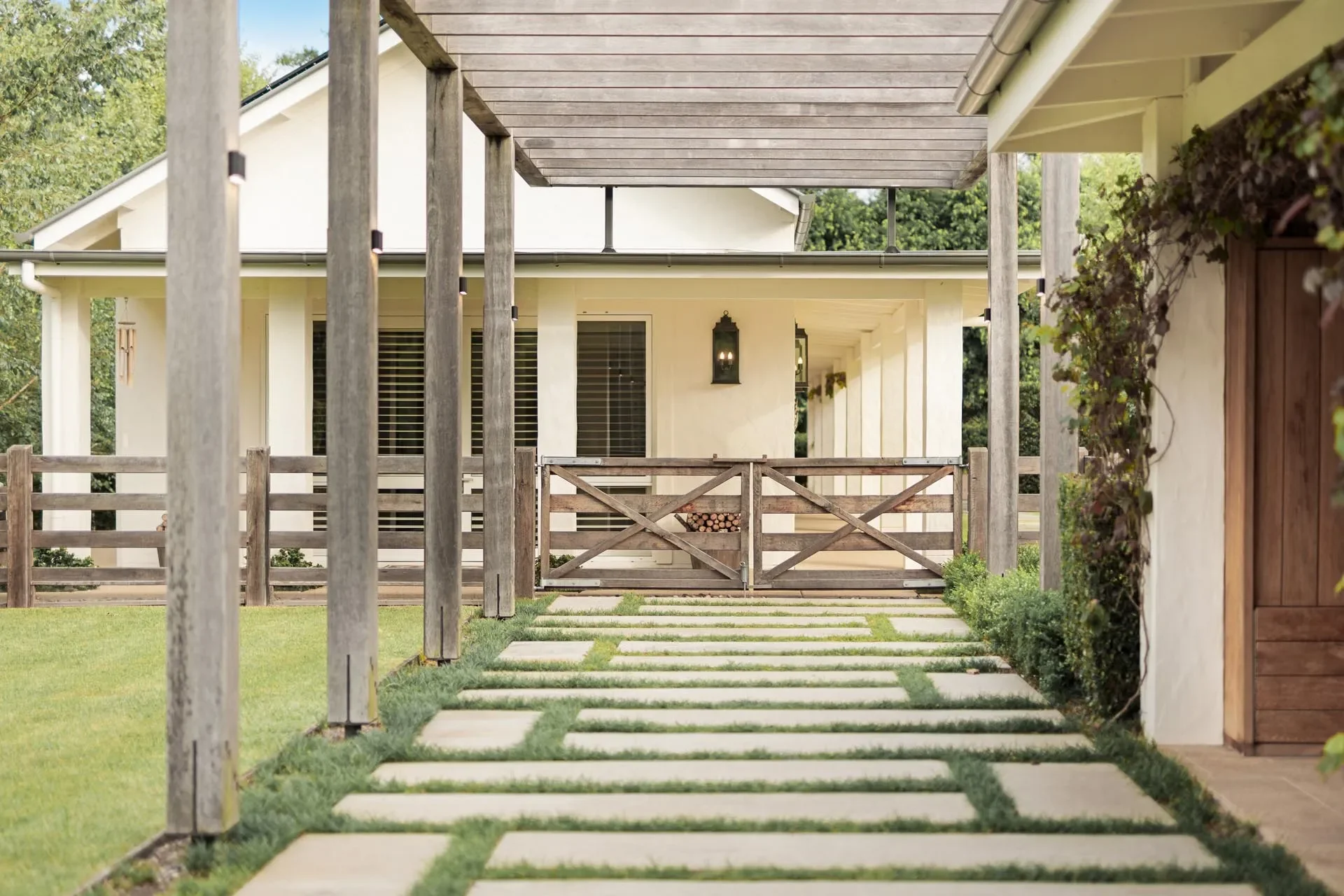 Front yard of a house with a wooden pathway leading to a porch, a wooden fence, and a white exterior wall with windows, lantern-style lights, and a wooden gate at the entrance.
