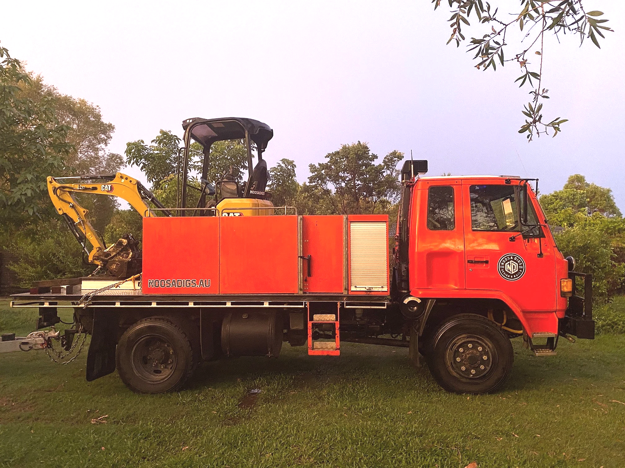 A red utility truck with a yellow excavator attachment parked on a grassy area, surrounded by trees under a cloudy sky.