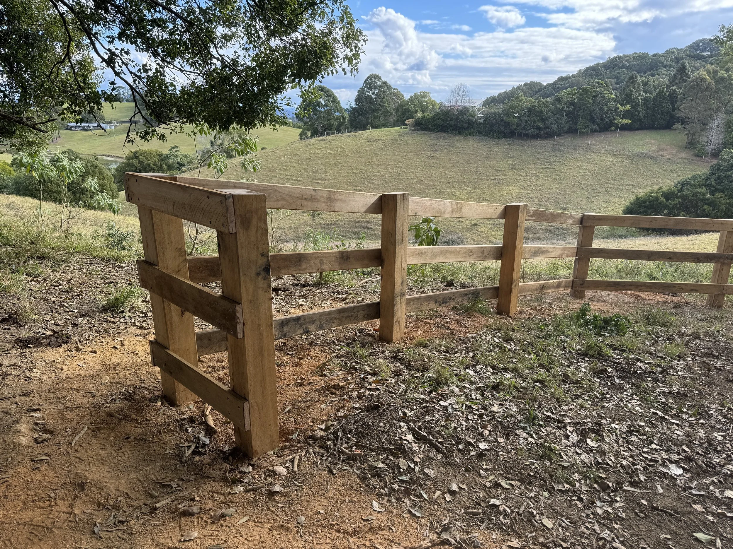 View of a green hilly landscape with trees, partly cloudy sky, and a wooden fence in the foreground.