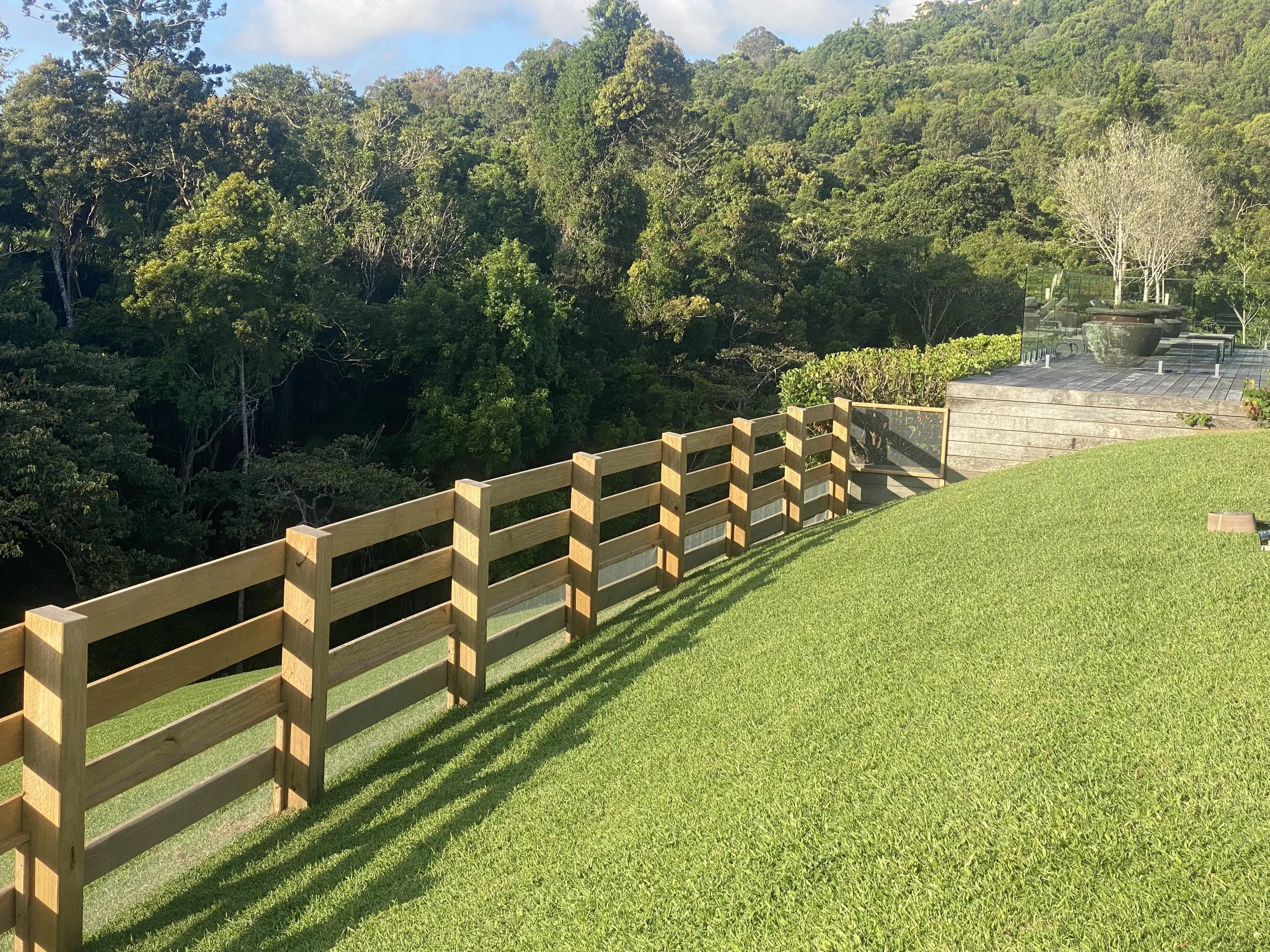A backyard with a wooden fence, lush green lawn, and a patio area with potted plants, trees, and forested hills in the background under a partly cloudy sky.