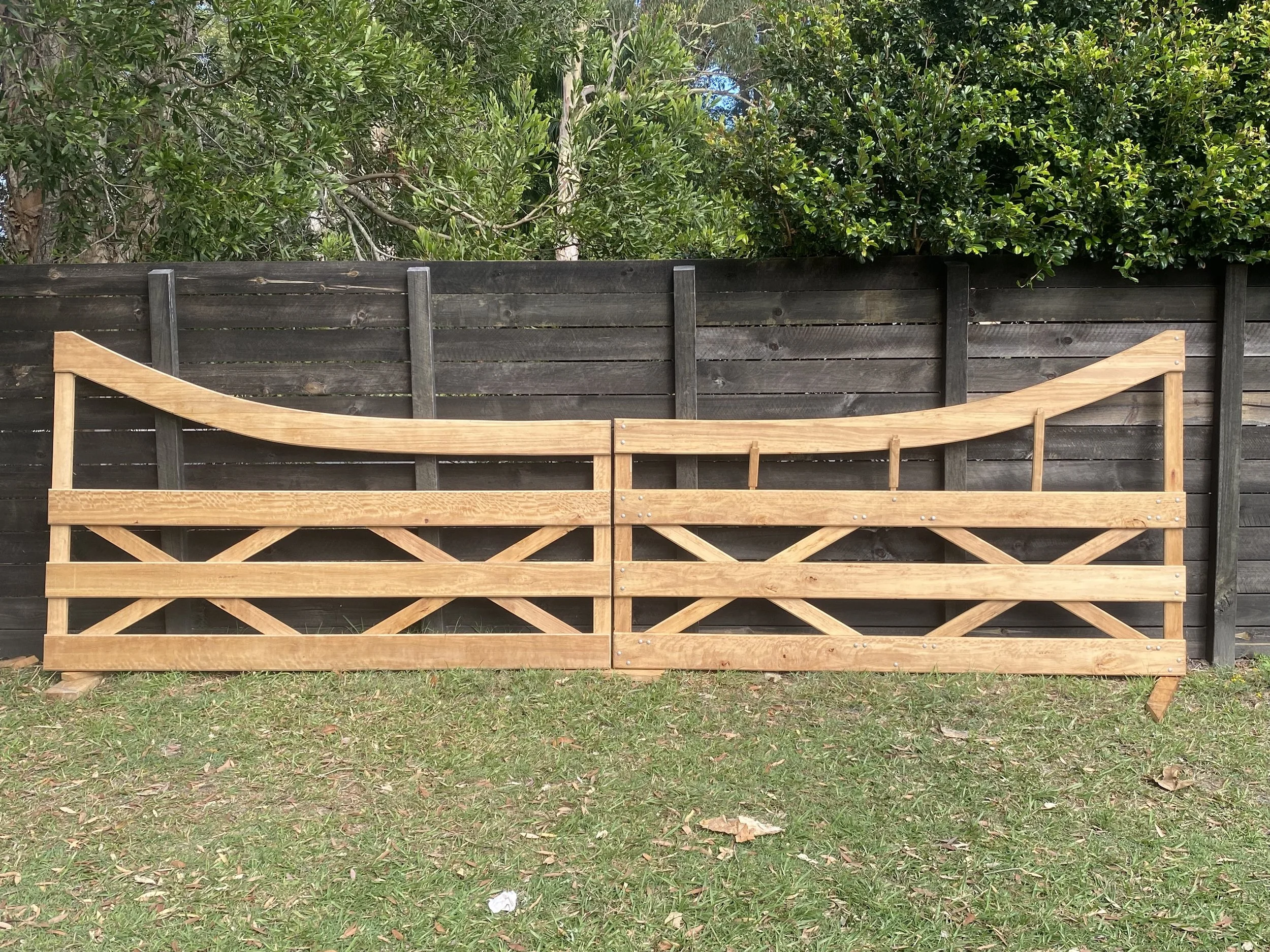 A partially assembled wooden bed frame in front of a black wooden fence with green shrubs and trees in the background.