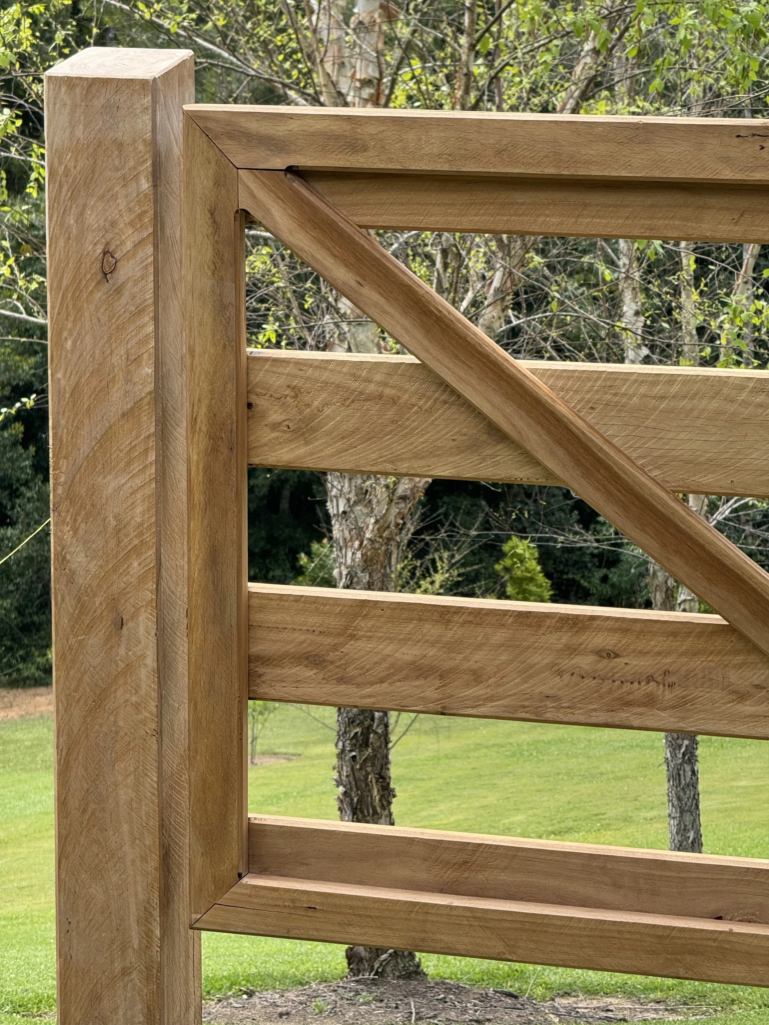 Close-up view of a wooden outdoor structure, possibly a fence or scaffolding, with a background of trees and grass.