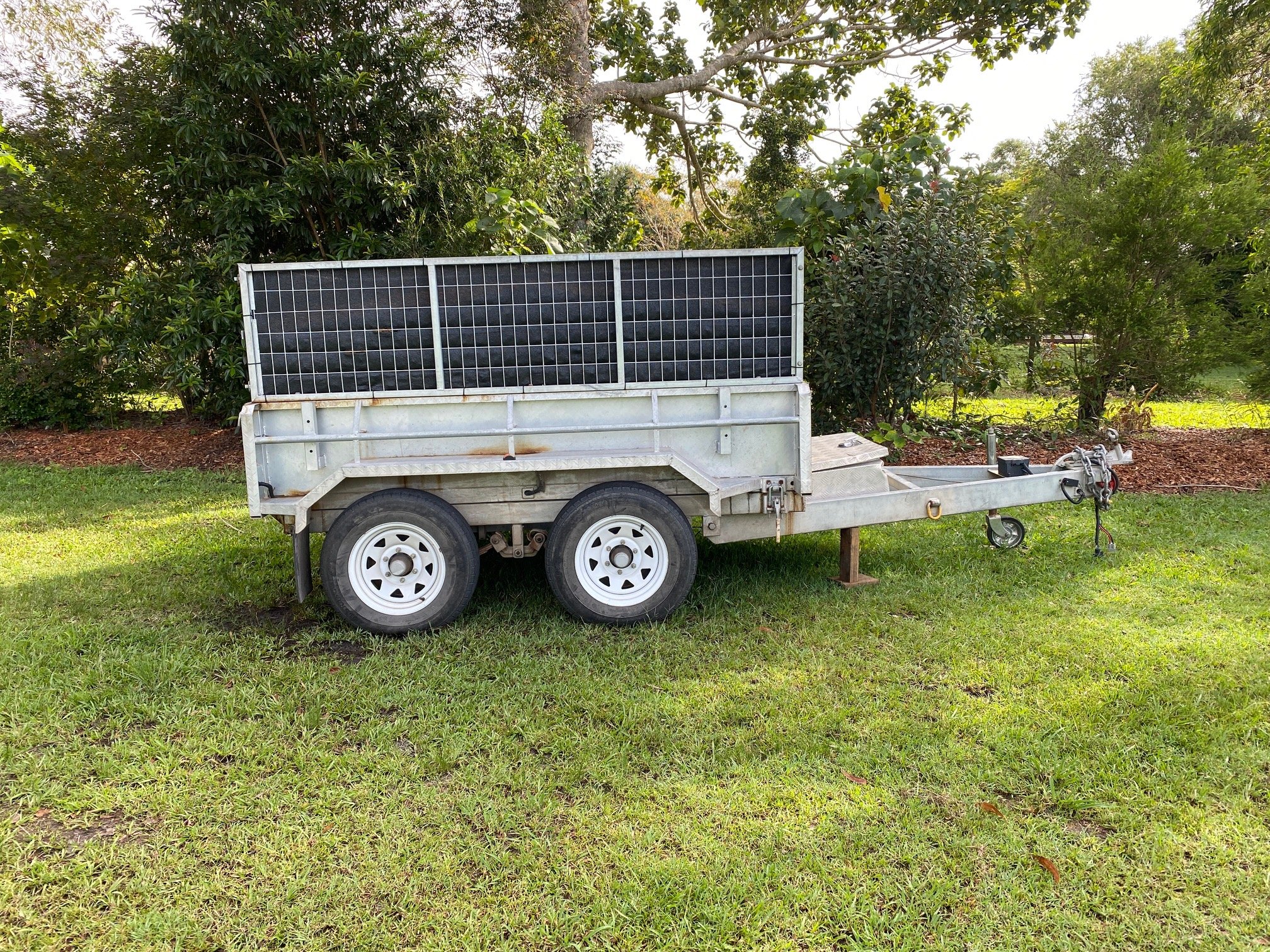 A trailer with solar panels on top, parked on grass, surrounded by trees and bushes.