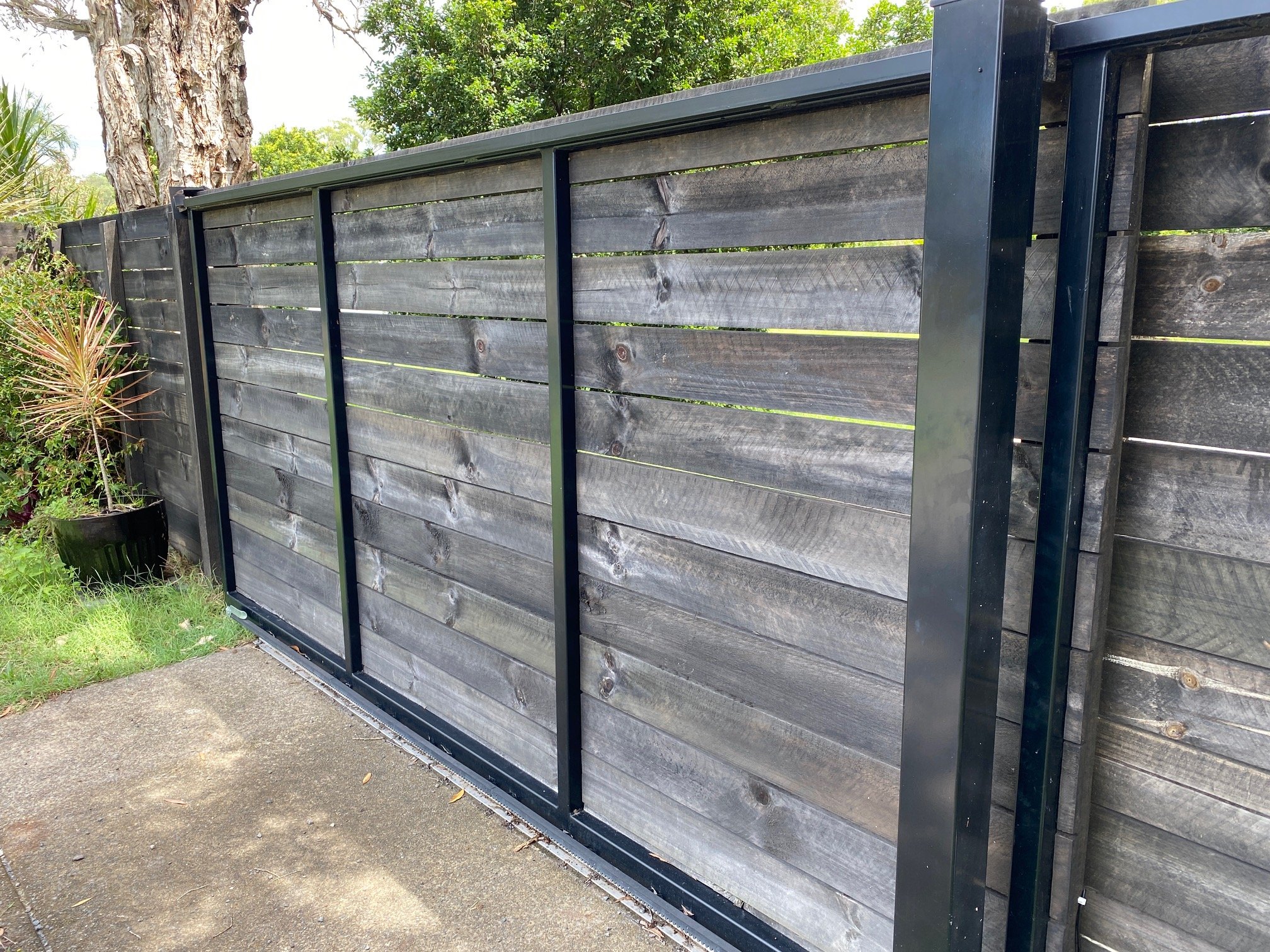 Black metal sliding gate with weathered wooden panels, set in a green outdoor area with trees and plants nearby.