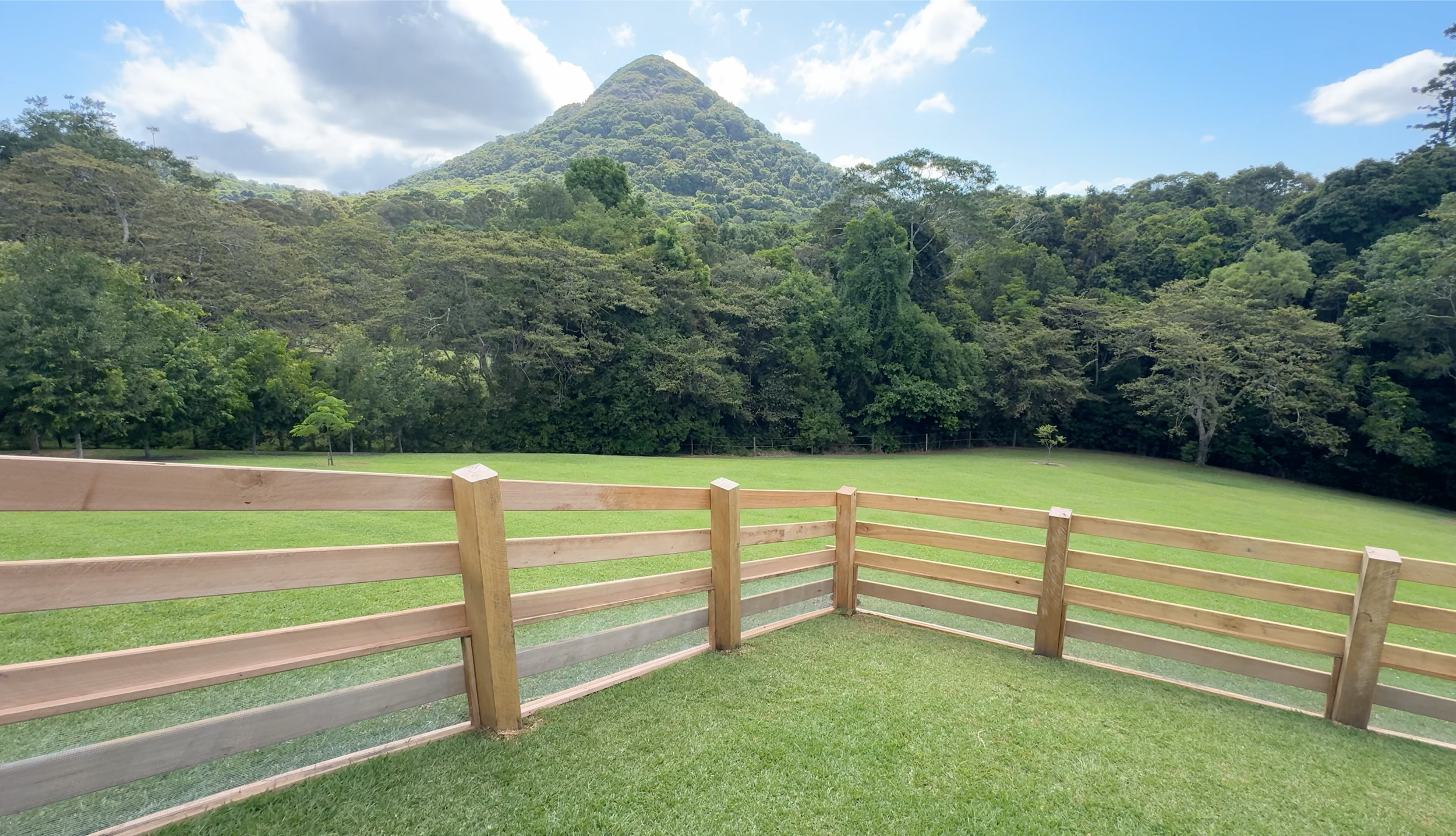A scenic view of a green mountain covered with trees overlooking a fenced grassy area with a wooden railing in the foreground, under a partly cloudy sky.