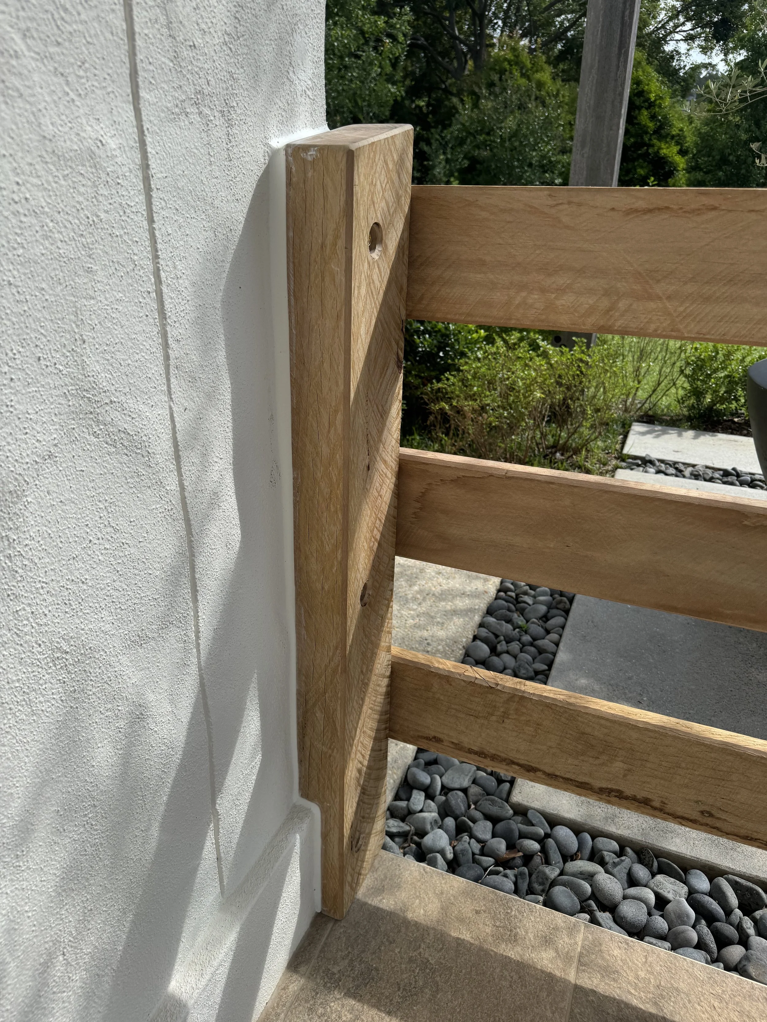 Close-up view of a wooden railing attached to a white stucco wall, with a background of greenery and a paved area with rocks.