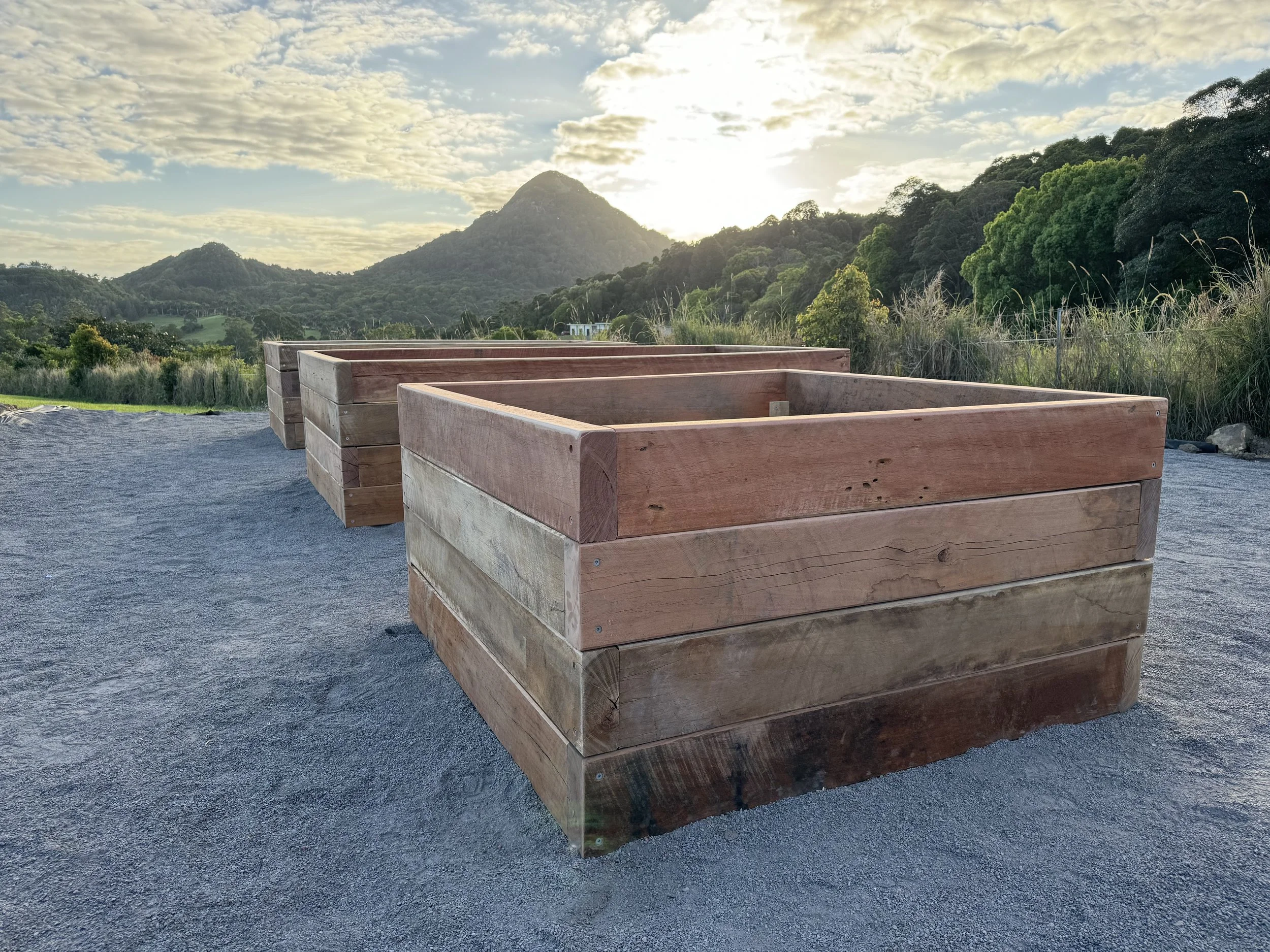 Three large wooden planters placed on a gravel surface outdoors, with a background of green trees and hills, and a partly cloudy sky during the daytime.