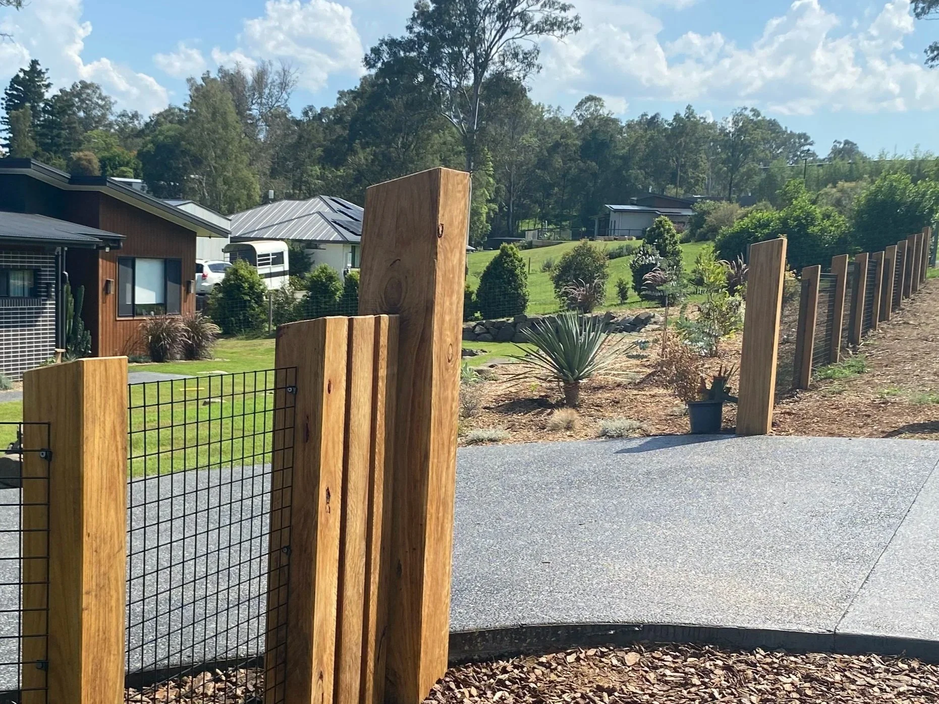 A wooden fence gate opening onto a paved driveway, with residential houses, trees, and bushes in the background under a partly cloudy sky.