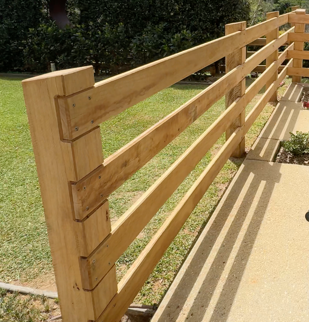 Wooden railing being installed along a sidewalk, with shadows cast on the ground.