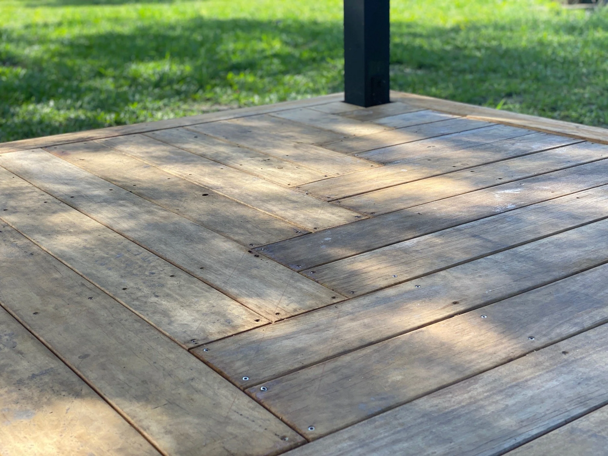 Close-up image of a wooden deck with visible nails, with a black metal post at the edge. The background shows blurred green grass and sunlight.