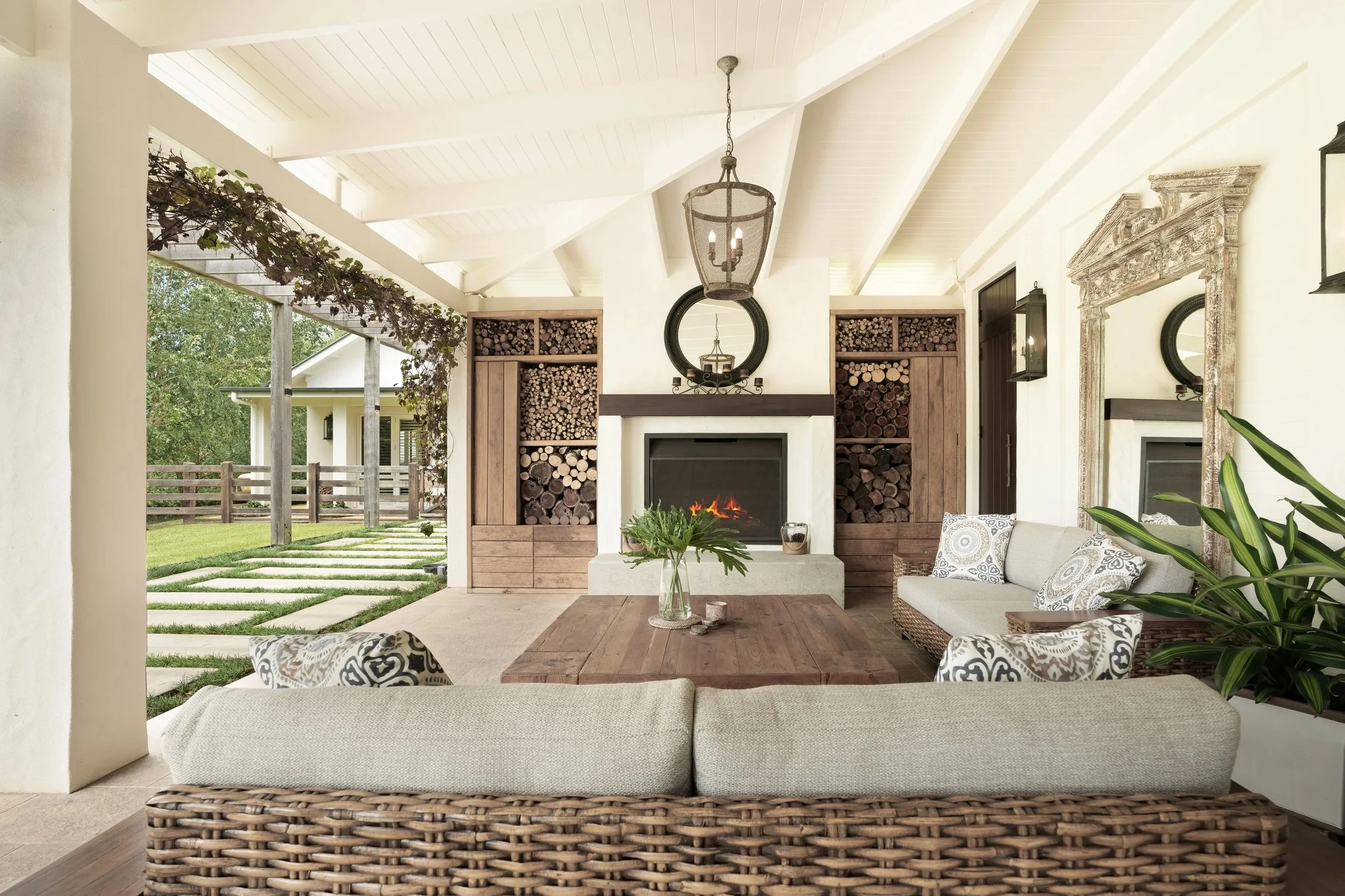 A cozy outdoor living space with a fireplace and built-in wood storage, featuring wicker furniture with patterned cushions, a glass vase with greenery on a wooden table, and surrounded by greenery and a wooden fence.