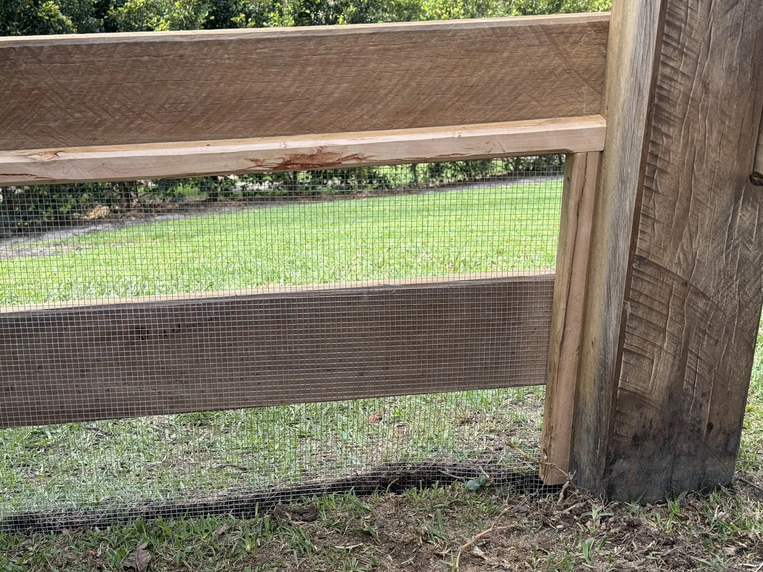 Wooden structure with wire mesh, outdoors on grass and soil, with trees in background.