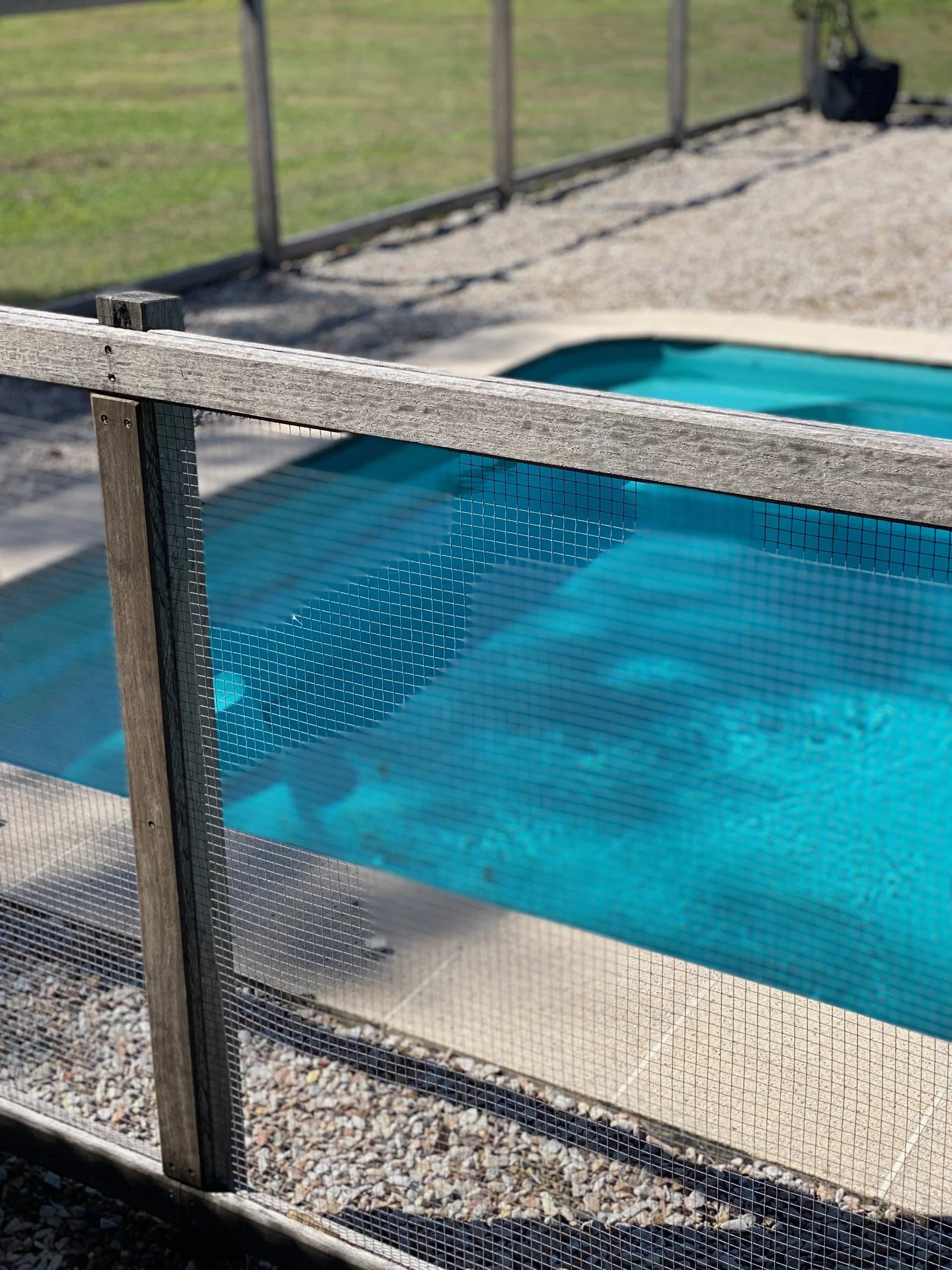 A small swimming pool with kidney shape filled with blue water, surrounded by gravel and enclosed by a wooden and wire fence.